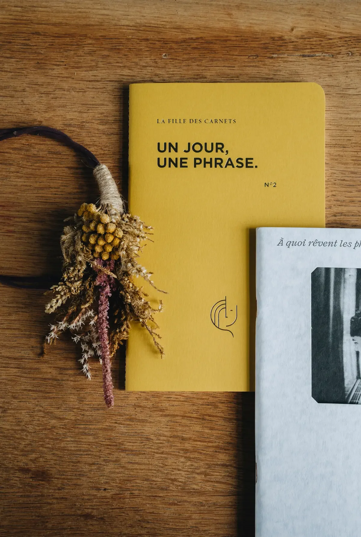 Livre jaune intitulé 'Un jour, une phrase' posé sur une table en bois, avec un bouquet de fleurs séchées attaché à une tête d'attache.