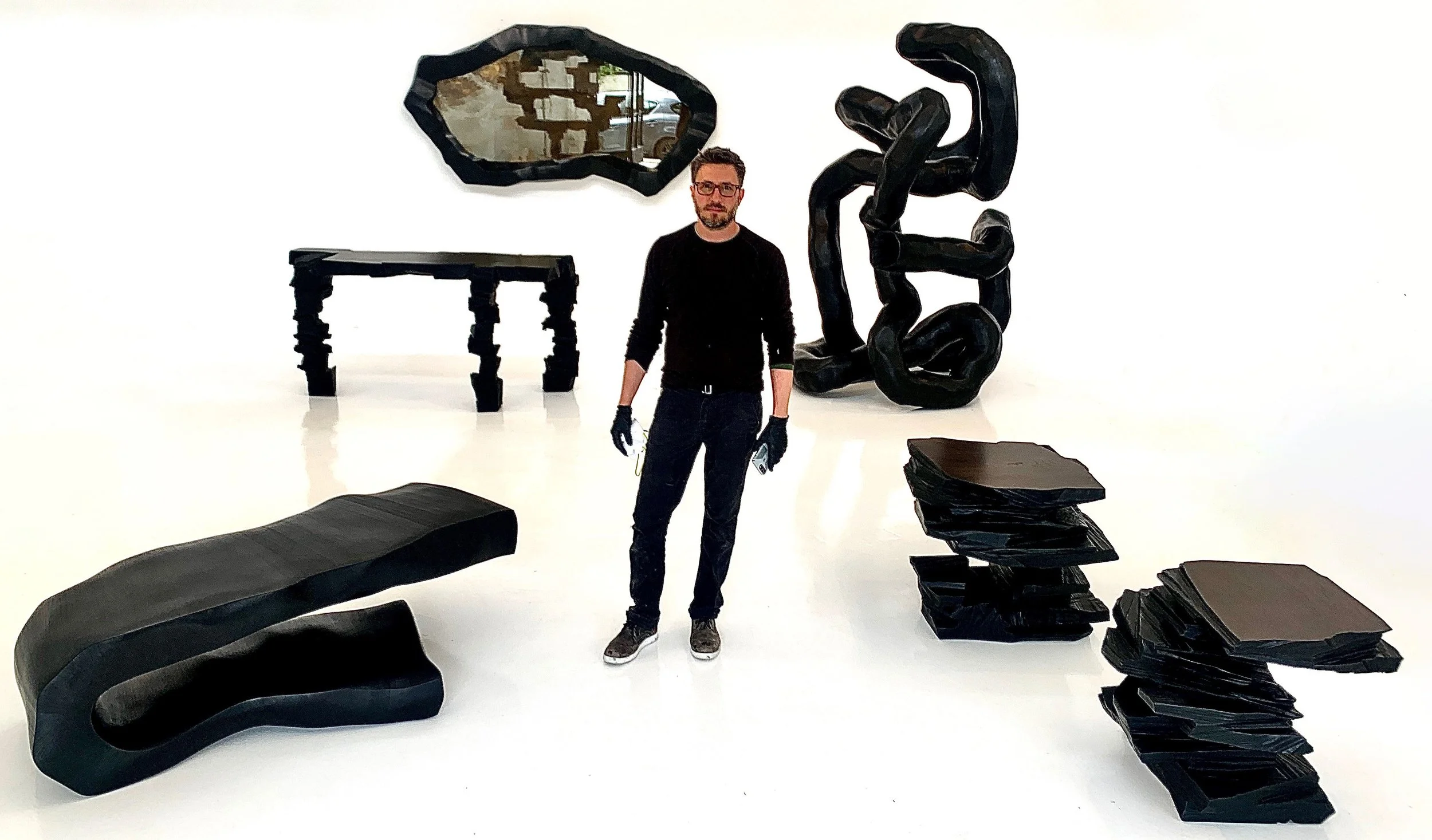 An art gallery installation featuring black sculptures, a mirror, and artist/designer Stefan Bishop standing in front of his artwork.