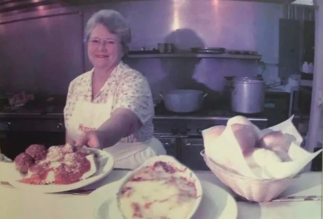 Smiling elderly woman in a kitchen holding a pizza on a tray with other pizzas and a basket of bread behind her.