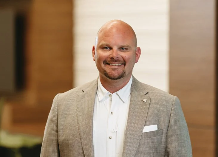 Bald man in beige suit smiling at camera, indoors with wooden and white background.