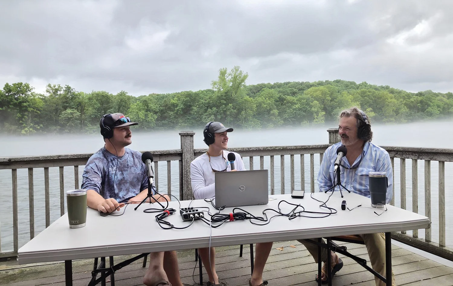 Three people sitting at a table outdoors by a body of water, recording a podcast. They are wearing headphones and microphones, with a laptop, phone, and drinks on the table. There is greenery and a misty lake in the background under cloudy skies.