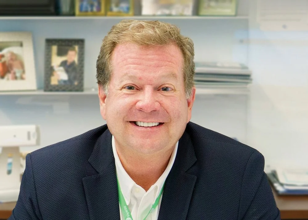 Smiling man in a suit sitting in an office with framed photos and shelves in the background.