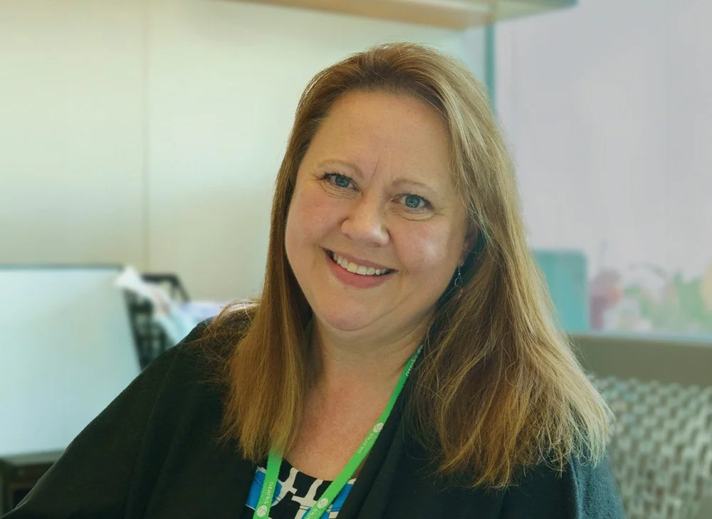A woman with shoulder-length light brown hair, wearing a black top and a green lanyard, smiling in an office environment.