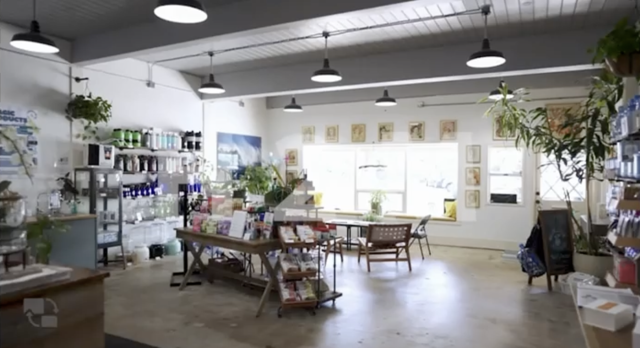 Interior of a retail store with shelves, tables, and seating area, filled with plants and various products under hanging lights, with large windows in the background.