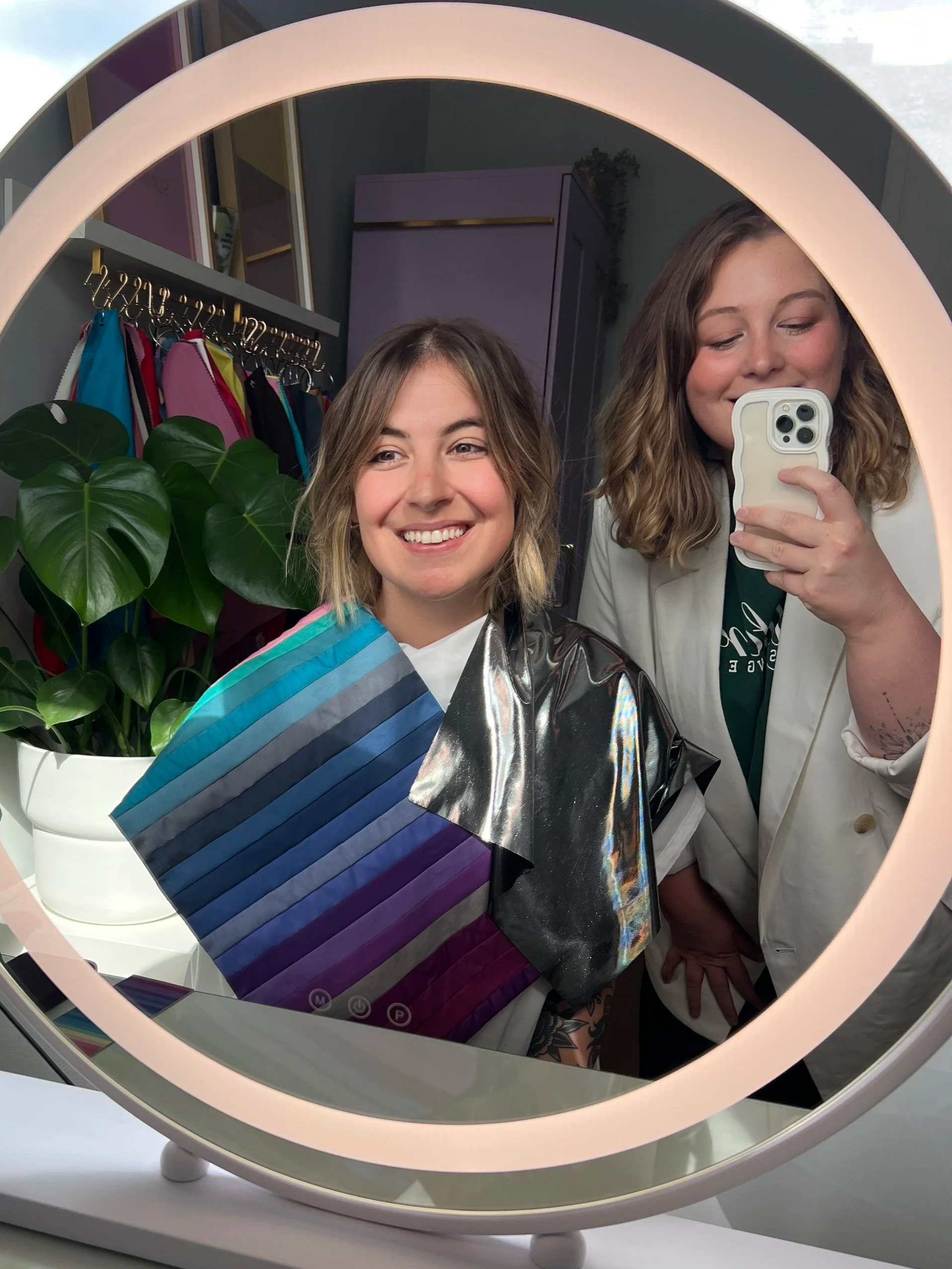 A woman with shoulder-length hair smiling at herself in a round mirror, taking a selfie with her phone. Behind her, a purple cabinet and colorful hanging backpacks are visible, along with large green houseplants.