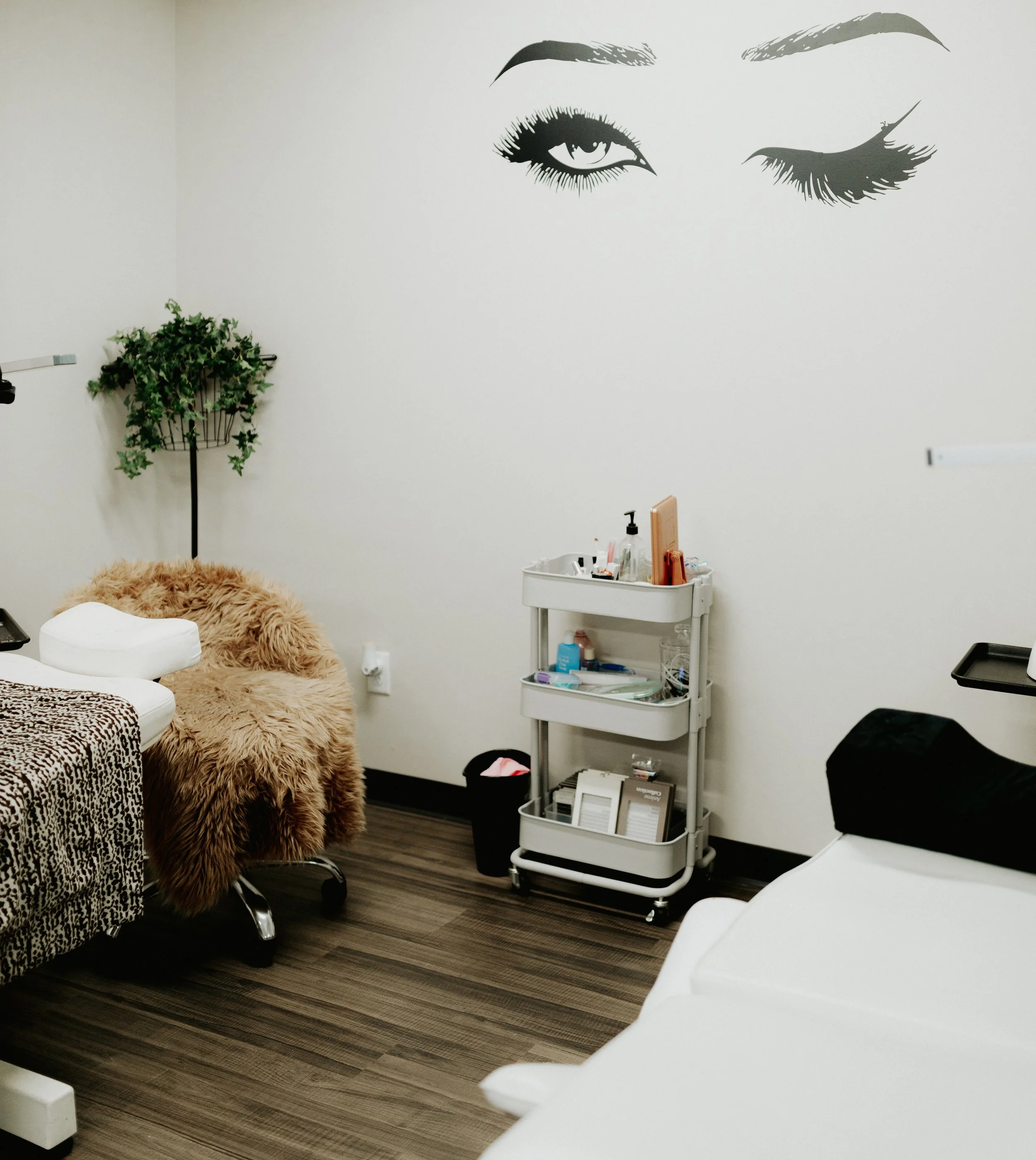 A room in a medical or beauty clinic with a wall art of a woman’s closed and open eyes, a white examination bed, a small rolling cart with skincare products, and a faux fur-covered stool all signifying lash technicians and lash extensions.