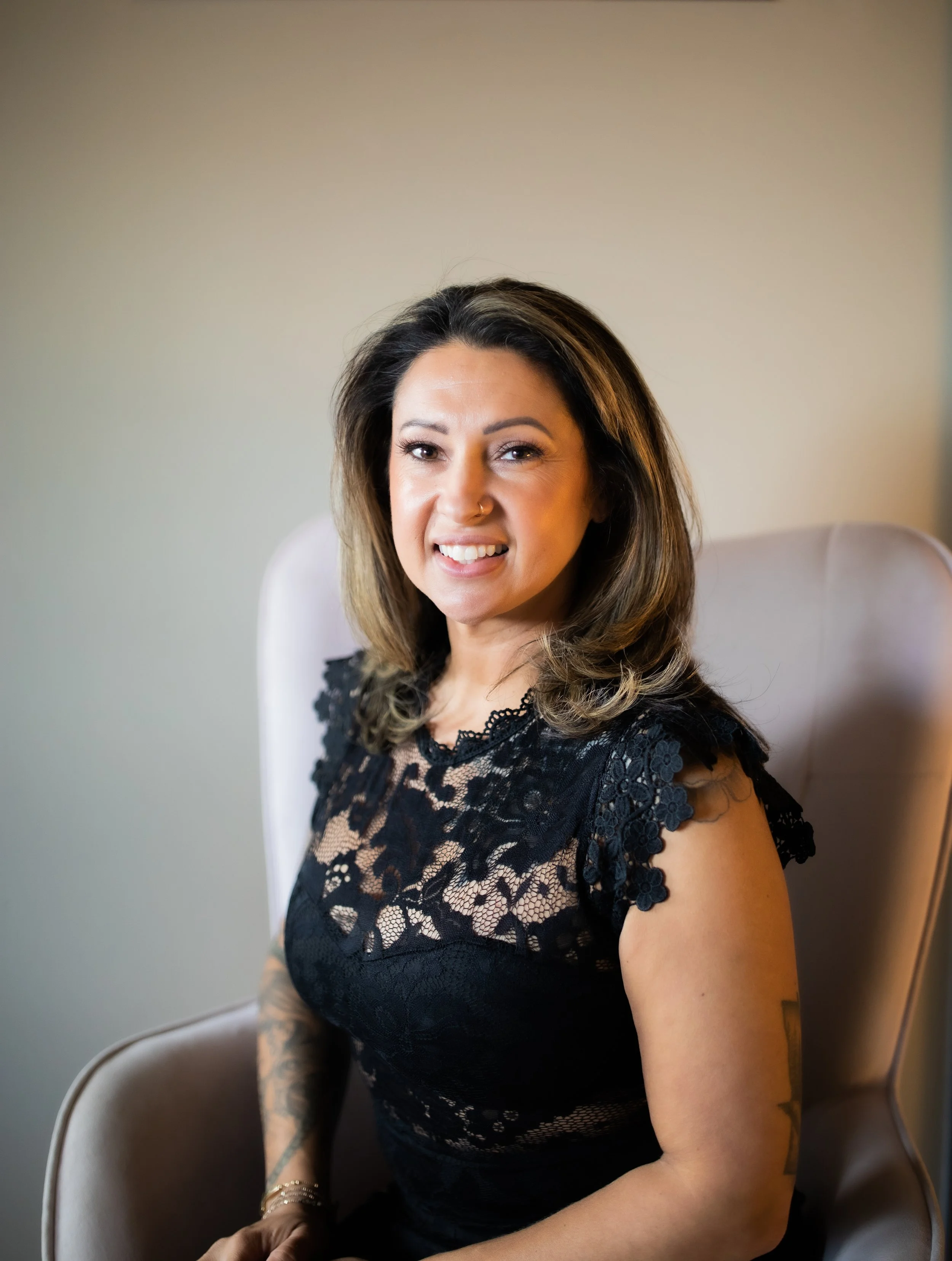 A salon owner woman with shoulder-length dark hair, sitting on a beige chair, wearing a black lace dress, smiling at the camera.
