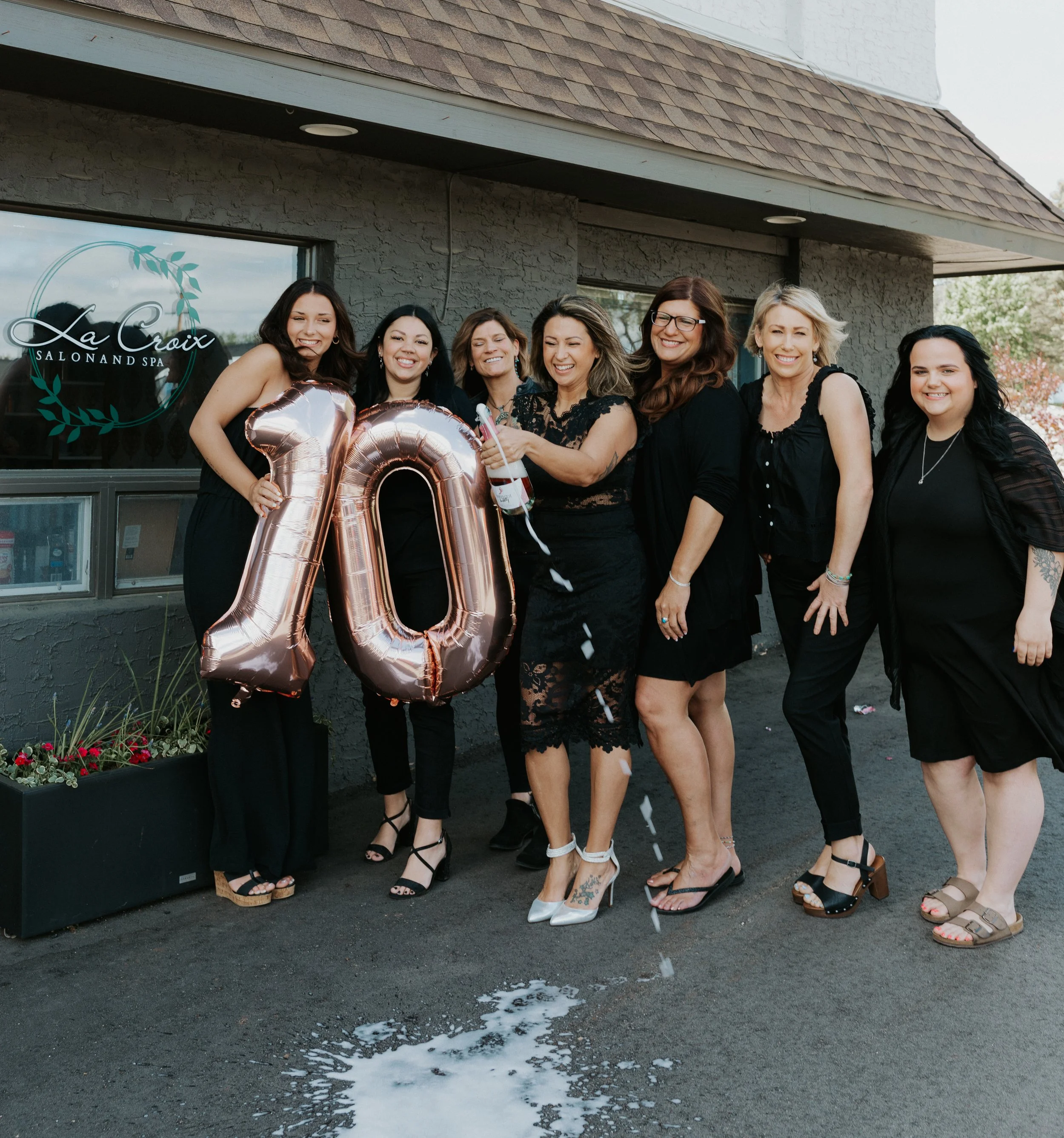 Group of women celebrating outside a salon, holding big rose gold balloons shaped as the number 10, with confetti falling.