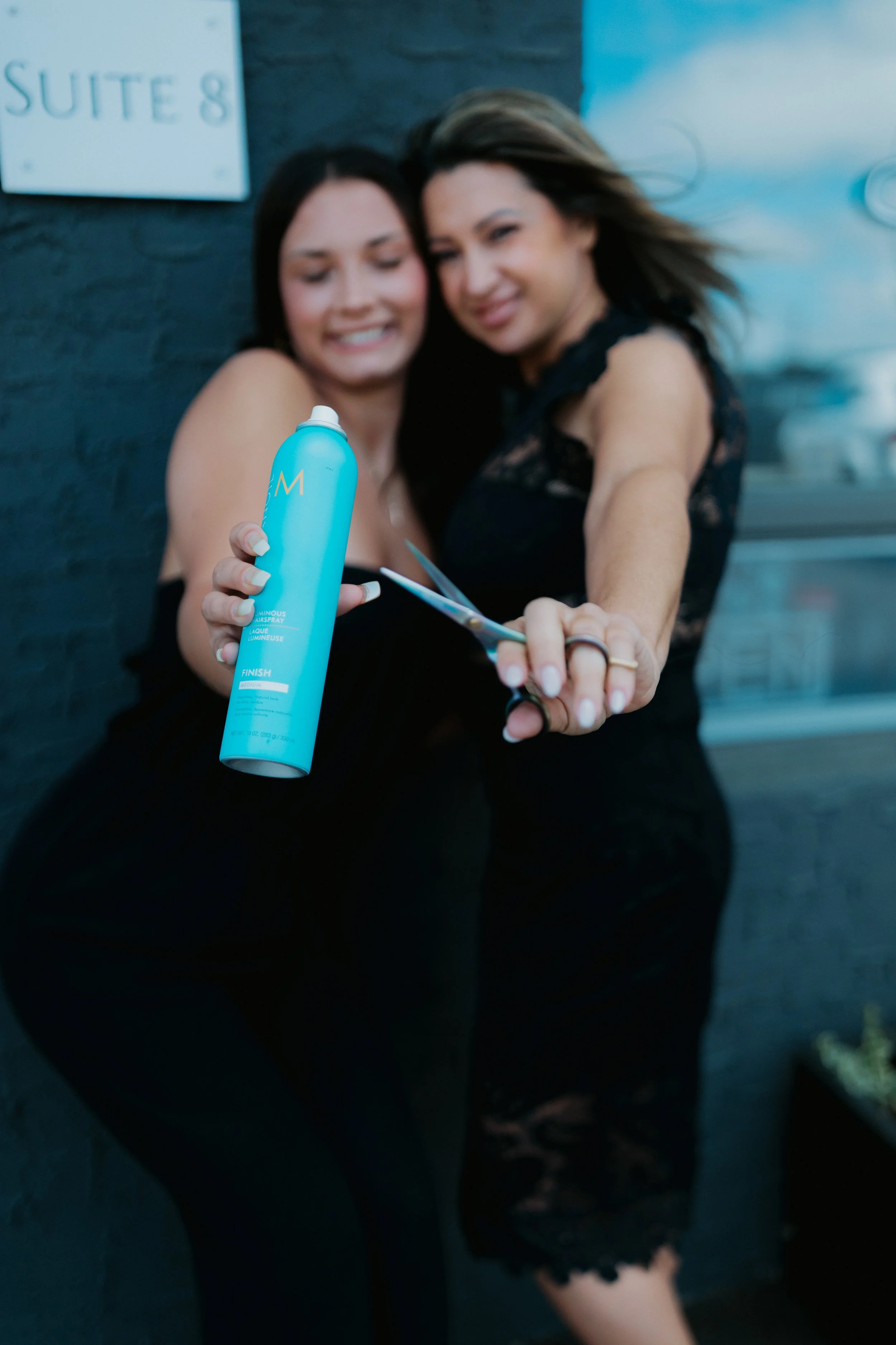 Two women in black dresses take a selfie together, with one holding a can of morrocan oil hair spray and a pair of sheers towards the camera.