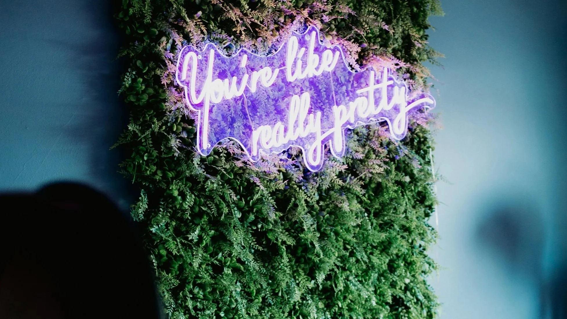 Neon sign with purple glow that reads "You're like really pretty," mounted on a wall adorned with a lush arrangement of two-tone purple and green plants.