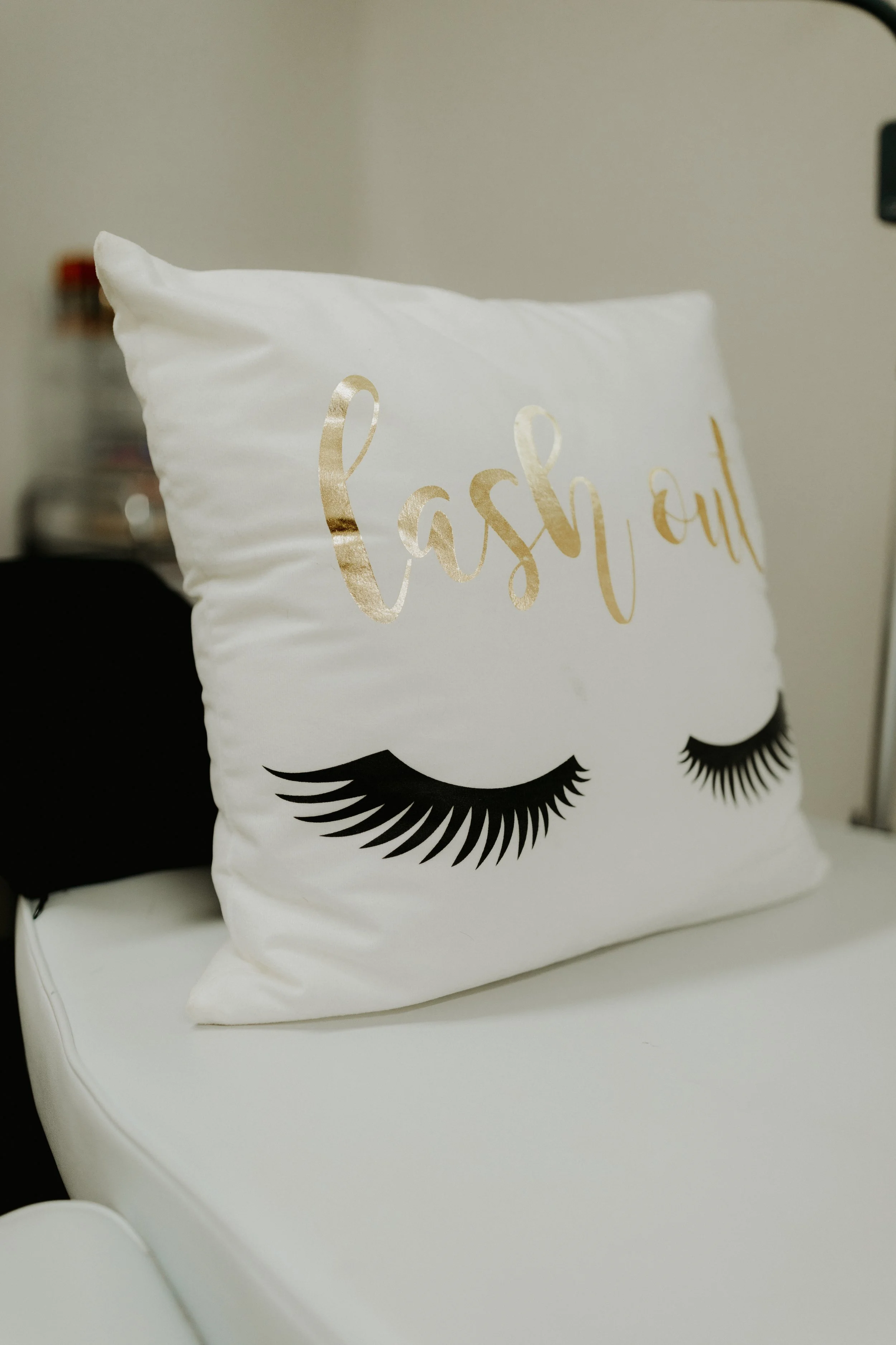 Decorative white pillow with gold cursive writing and black eyelash graphics, resting on a white surface.