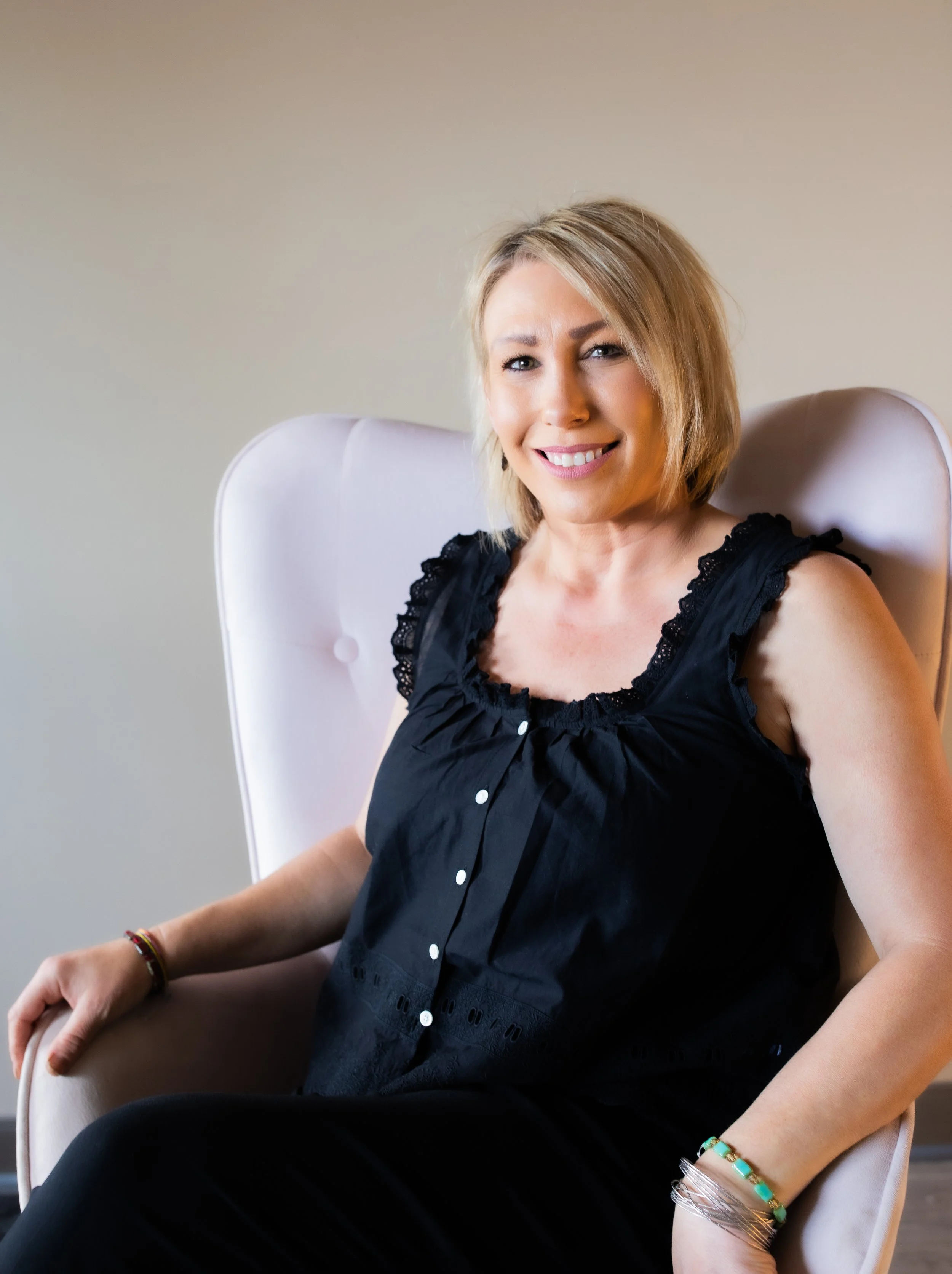 A stylist with blonde hair sitting in a white chair, smiling and looking at the camera. She is wearing a black sleeveless top with lace details and black pants.