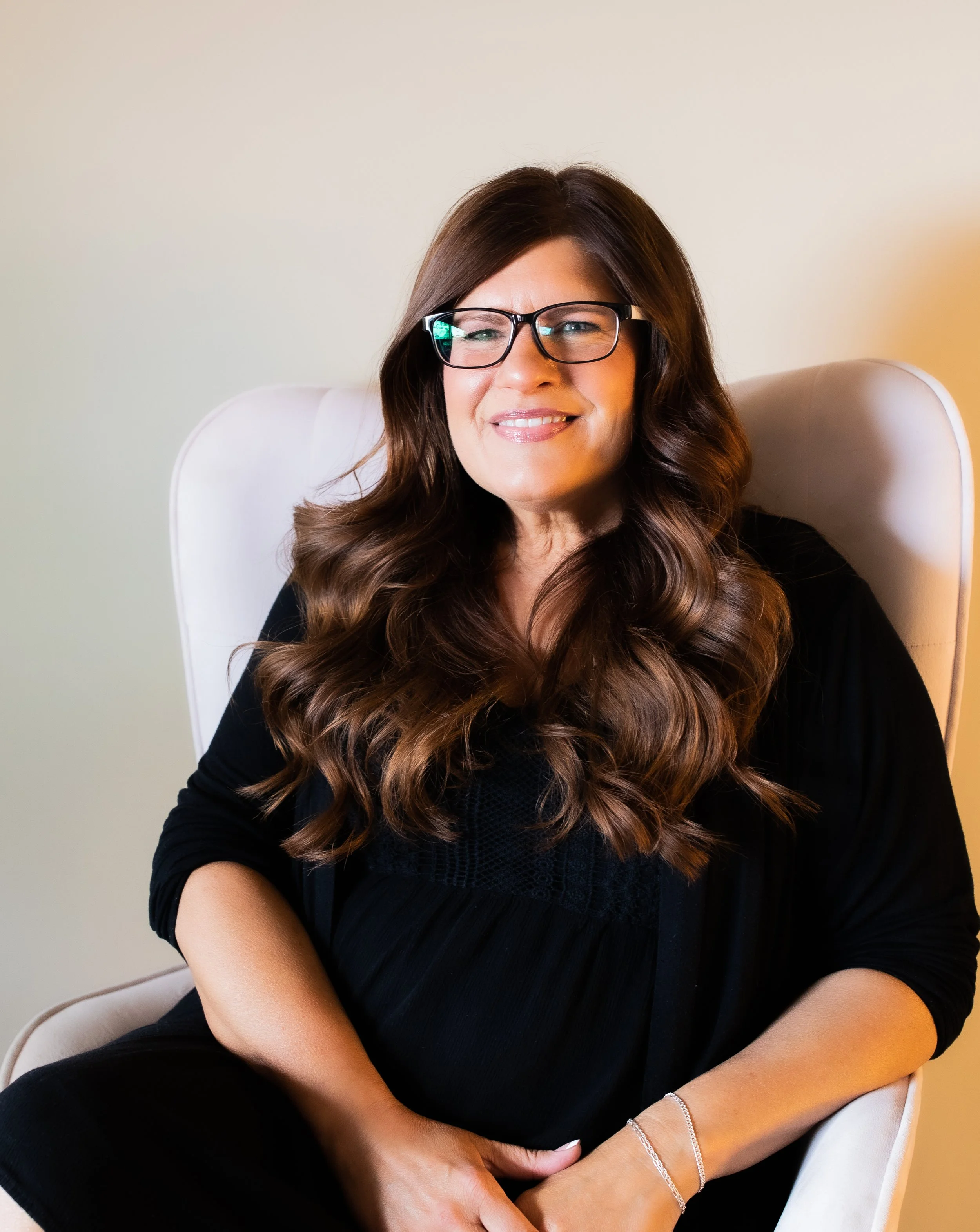 A stylist with long wavy brown hair, wearing glasses and a black top, sitting on a white armchair, smiling at the camera.