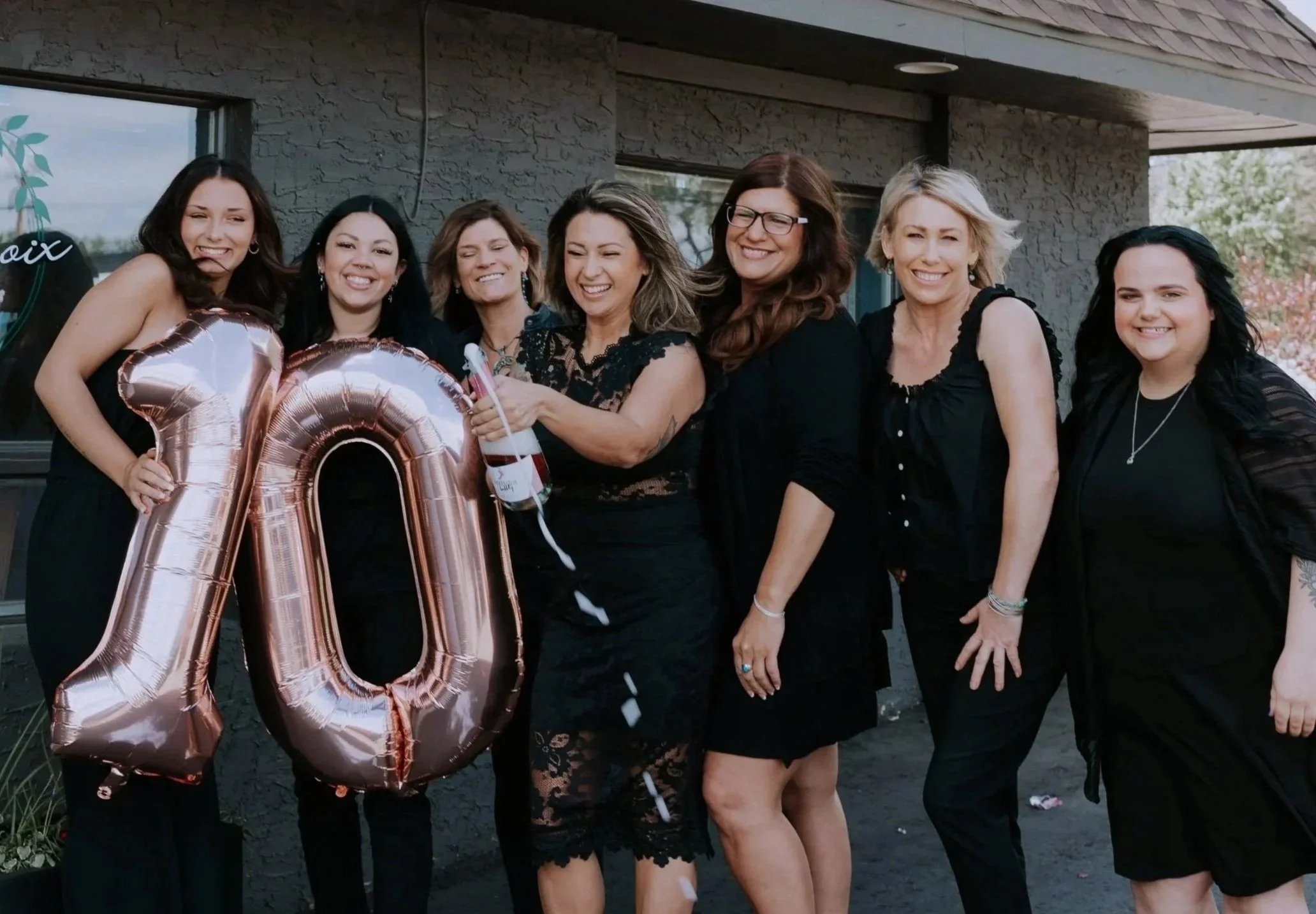 Group of seven hair stylists celebrating an anniversary outside a salon building, with a large rose gold balloon shaped as the number 10, dressed in black, smiling and holding a bottle of champagne.