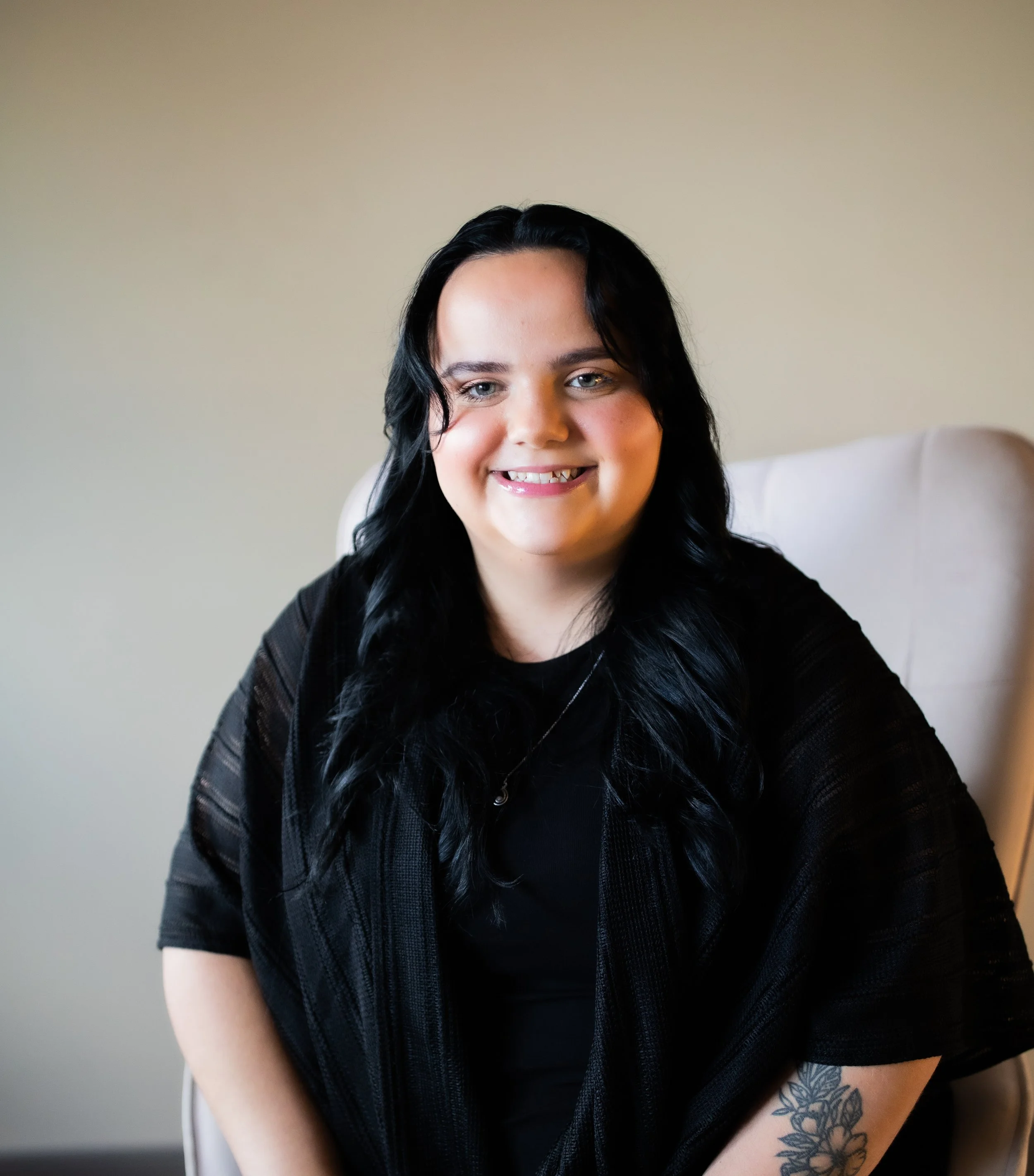 A hair color stylist with long black hair, smiling, sitting on a light-colored chair, wearing a black top and a black shawl, with a floral tattoo on her left forearm, in front of a plain beige wall.