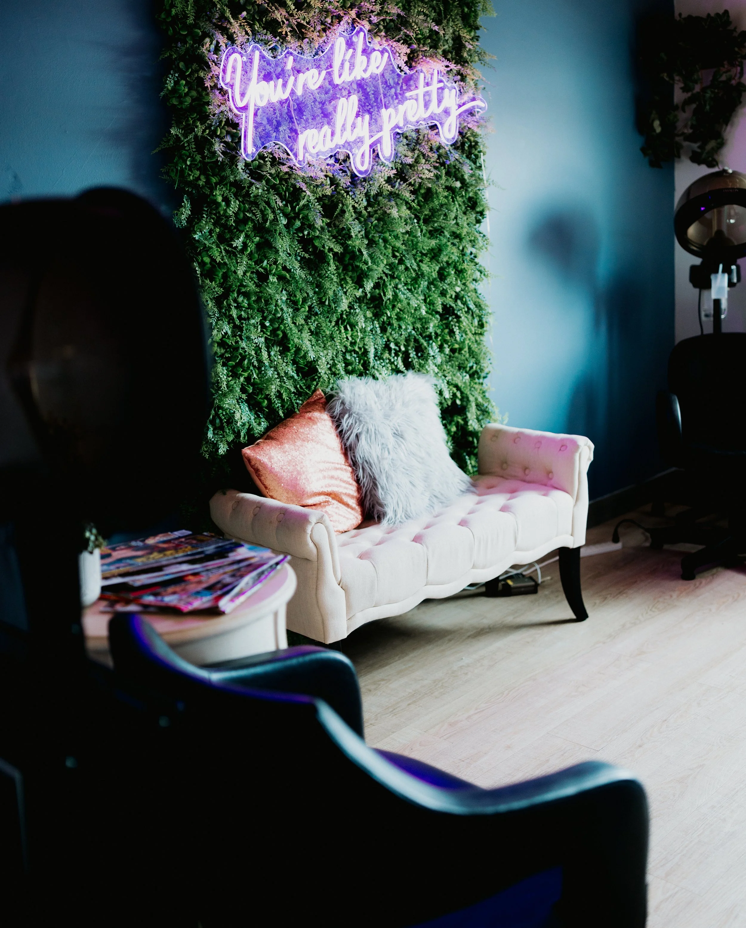 A cozy room with a pink tufted sofa accented with fluffy and metallic pillows, against a green foliage backdrop with a neon sign that says 'You're like really pretty'. There is a side table with magazines and a black chair with salon equipment nearby