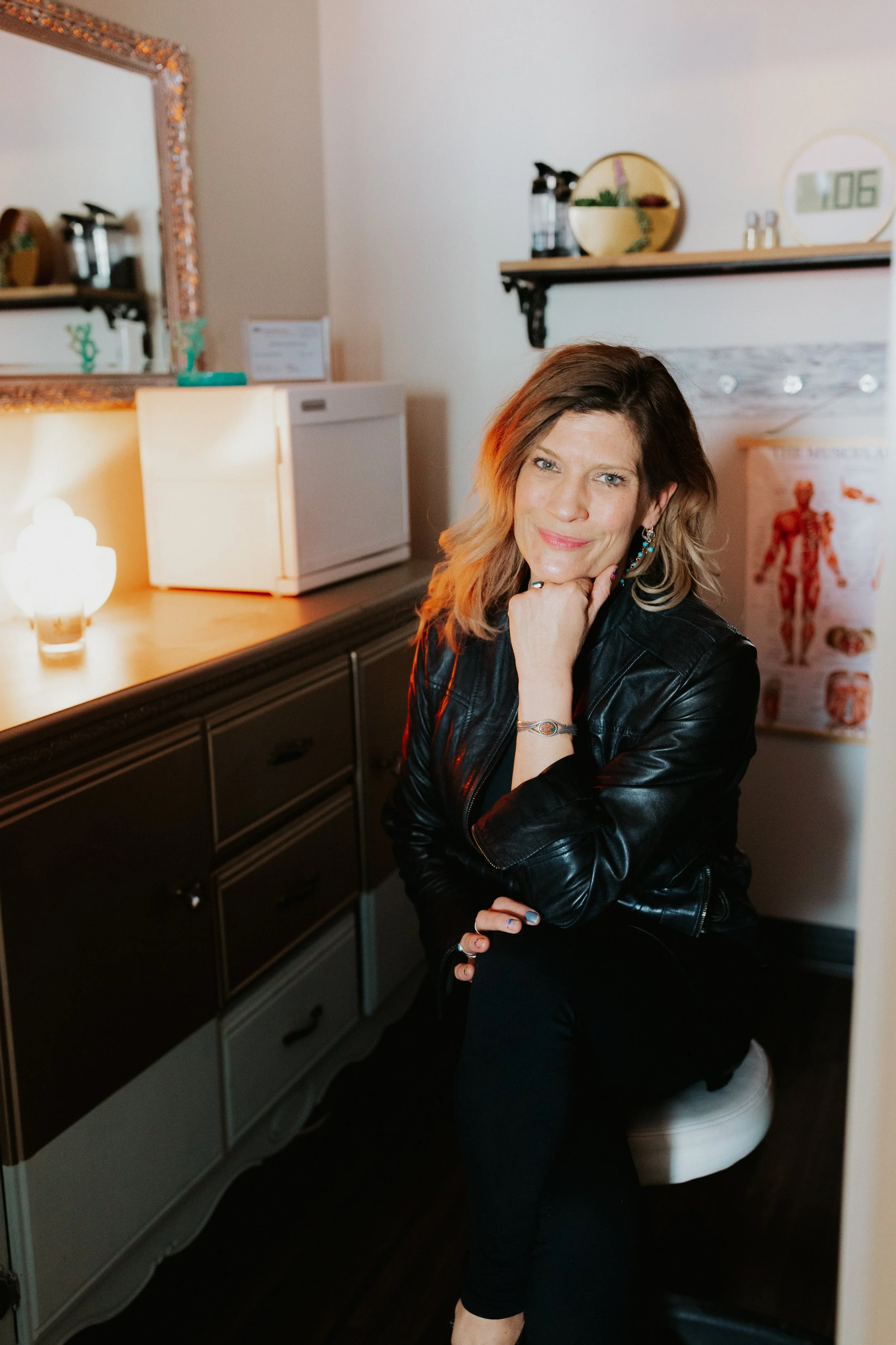 A woman with shoulder-length wavy hair, wearing a black leather jacket and black pants, is sitting on a toilet in a medical or wellness office. She is smiling slightly and resting her chin on her right hand.