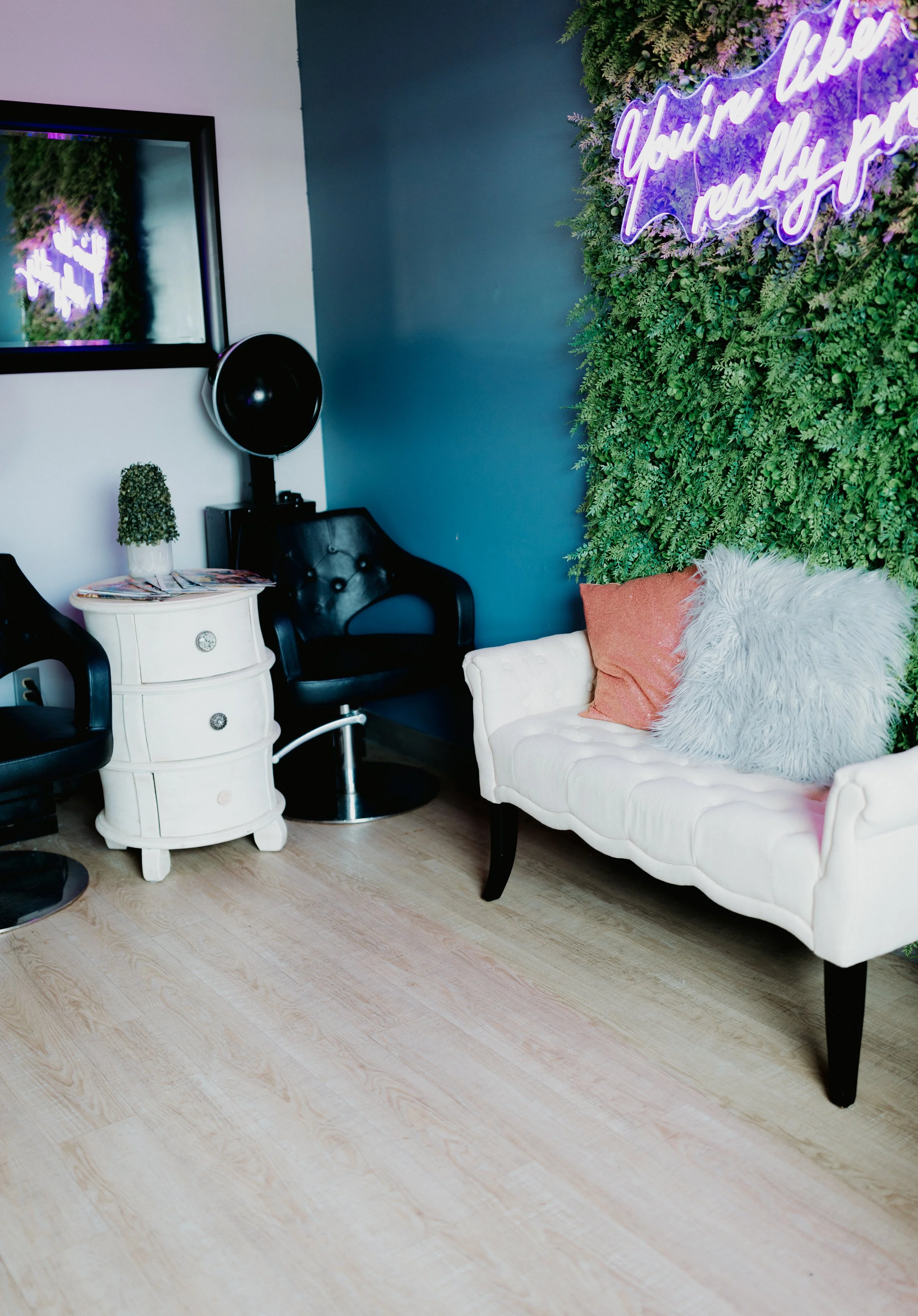 A salon or lounge area with a white tufted loveseat adorned with pink and fluffy white pillows, greenery on the wall behind it, a neon sign that reads 'You're like really pretty', two black salon chairs, a white small side table with magazines and a 