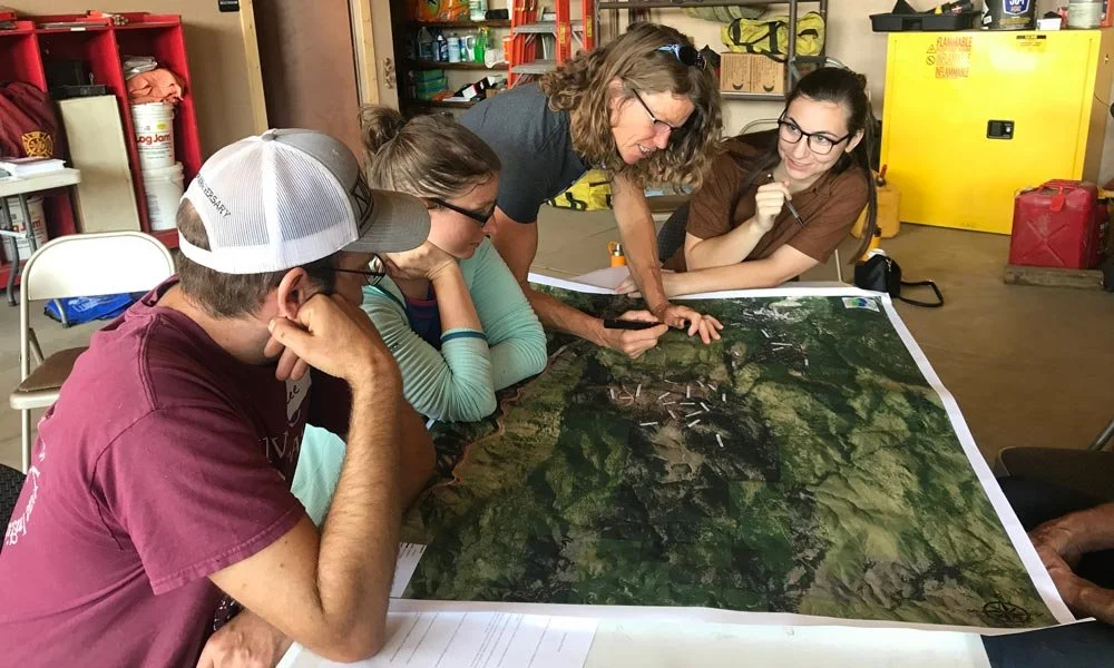 A group of five people gathered around a large map on a table, discussing and pointing at it in an indoor setting, likely a workshop or classroom.