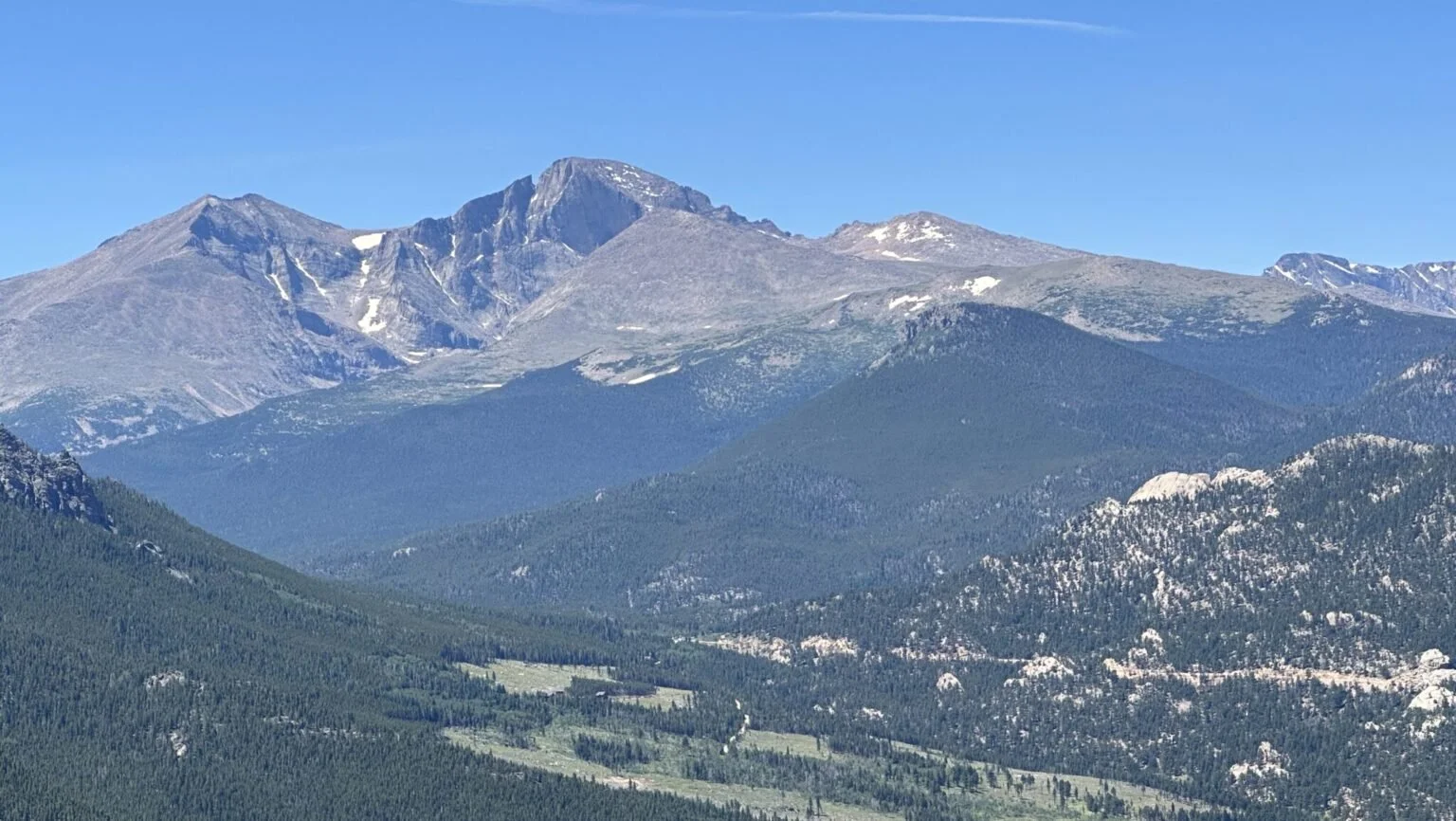 Scenic view of a mountain range with peaks, some snow patches, and forested slopes under a clear blue sky.