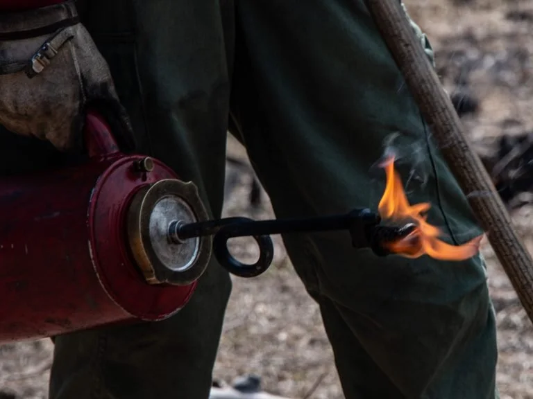 A person wearing dark green work clothes holding a red gasoline container with a long nozzle, igniting the fuel with a flame at the tip of the nozzle.