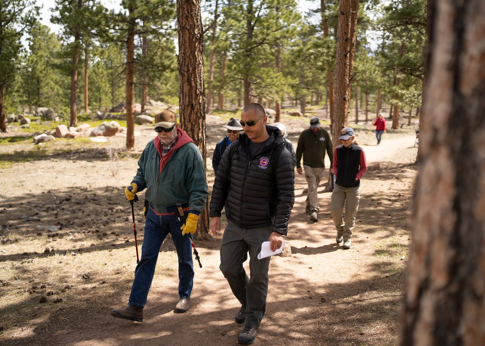 Group of people hiking through a forest, wearing outdoor gear and sunglasses, with trees and a dirt trail in the background.