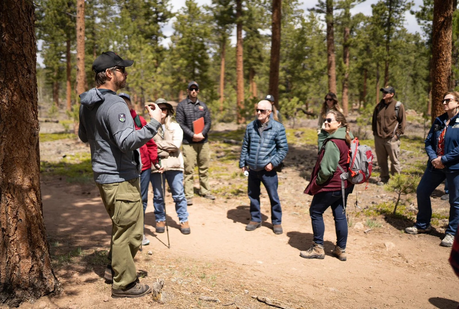 A man giving a guided tour to a group of people in a forest with tall pine trees, some people wearing backpacks and sunglasses, and a dirt trail underfoot.