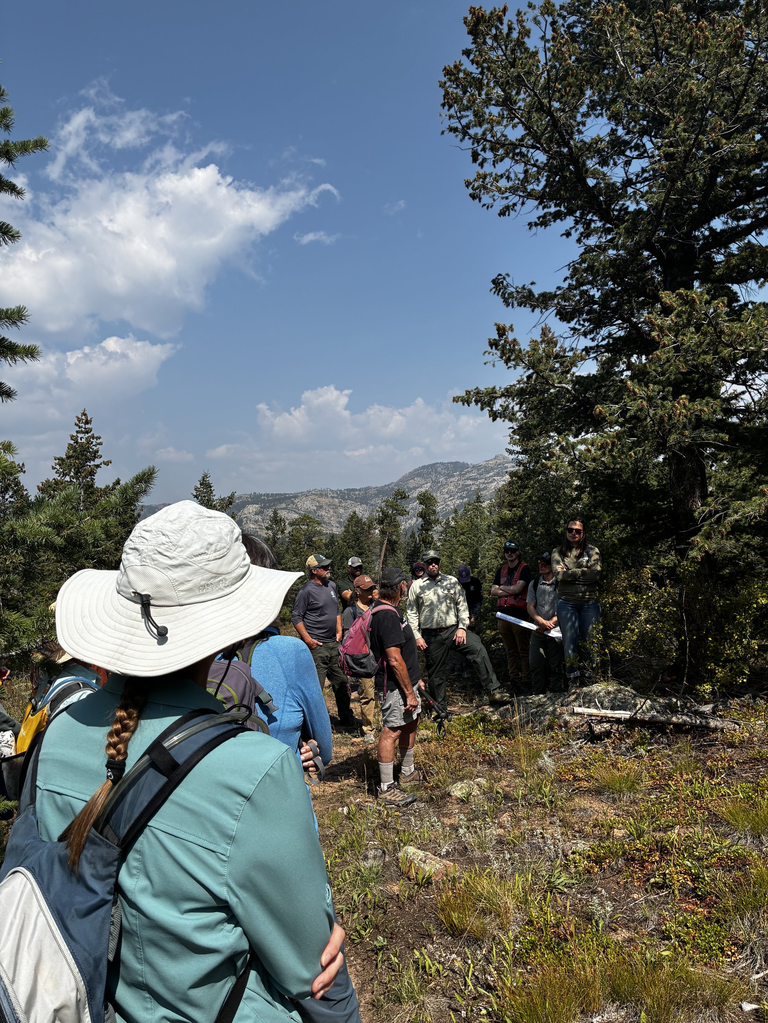 A group of hikers gathered outdoors in a forested area under a partly cloudy sky, with mountain terrain in the background. One person in the foreground is wearing a wide-brimmed white hat and a teal jacket, with a backpack. Other hikers are listening to a woman speaker, some wearing hats and sunglasses.