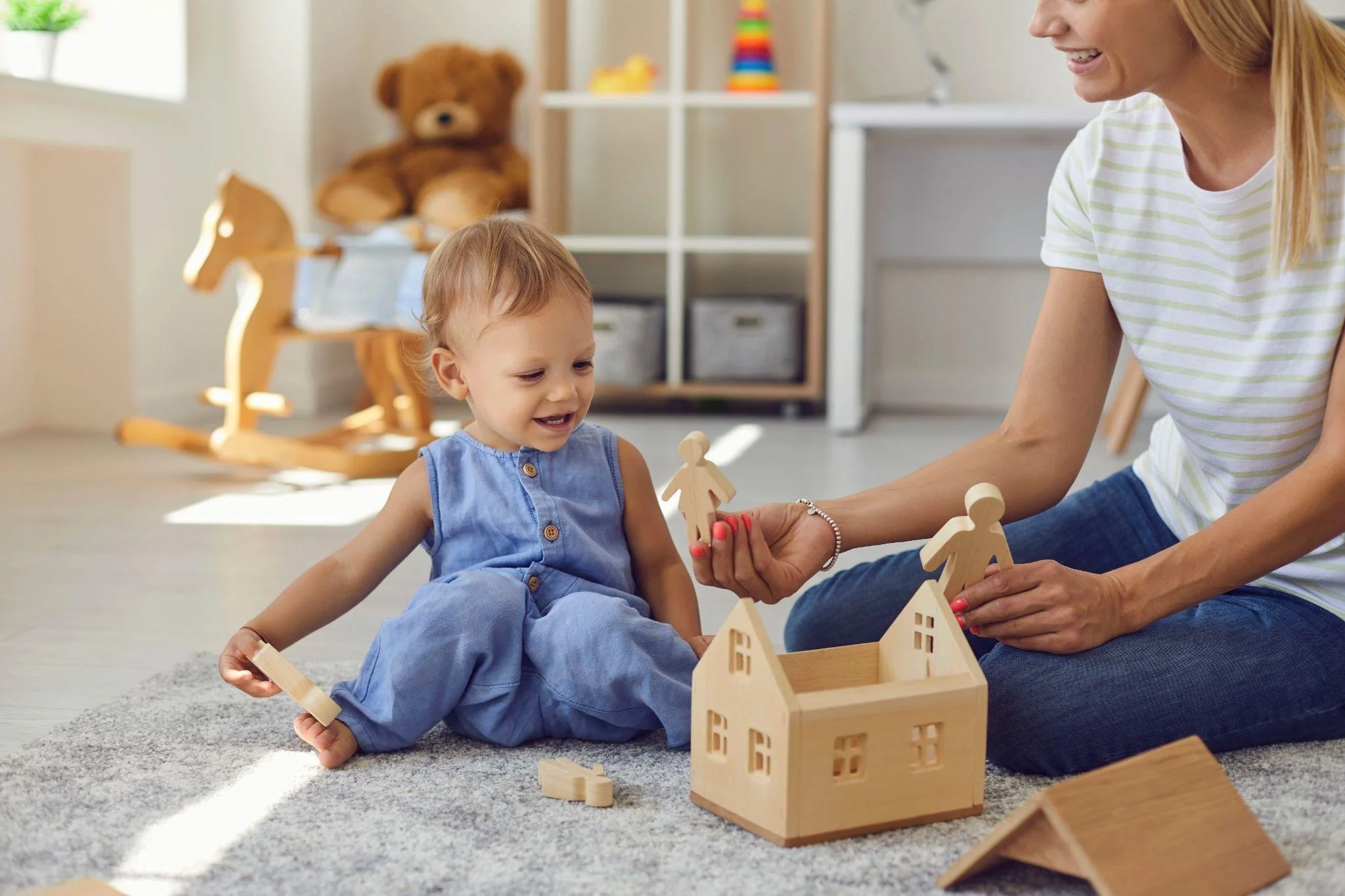 A young child and an adult woman sitting on a carpeted floor playing with wooden house and figurines toys, in a room with a rocking horse, a teddy bear, and storage shelves.