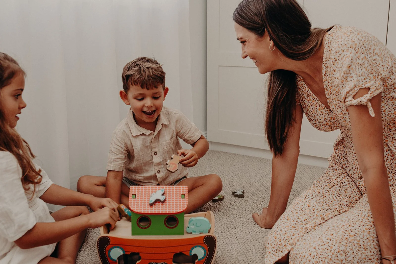 A woman and two children sitting on the floor playing with a wooden toy with animals and blocks inside.