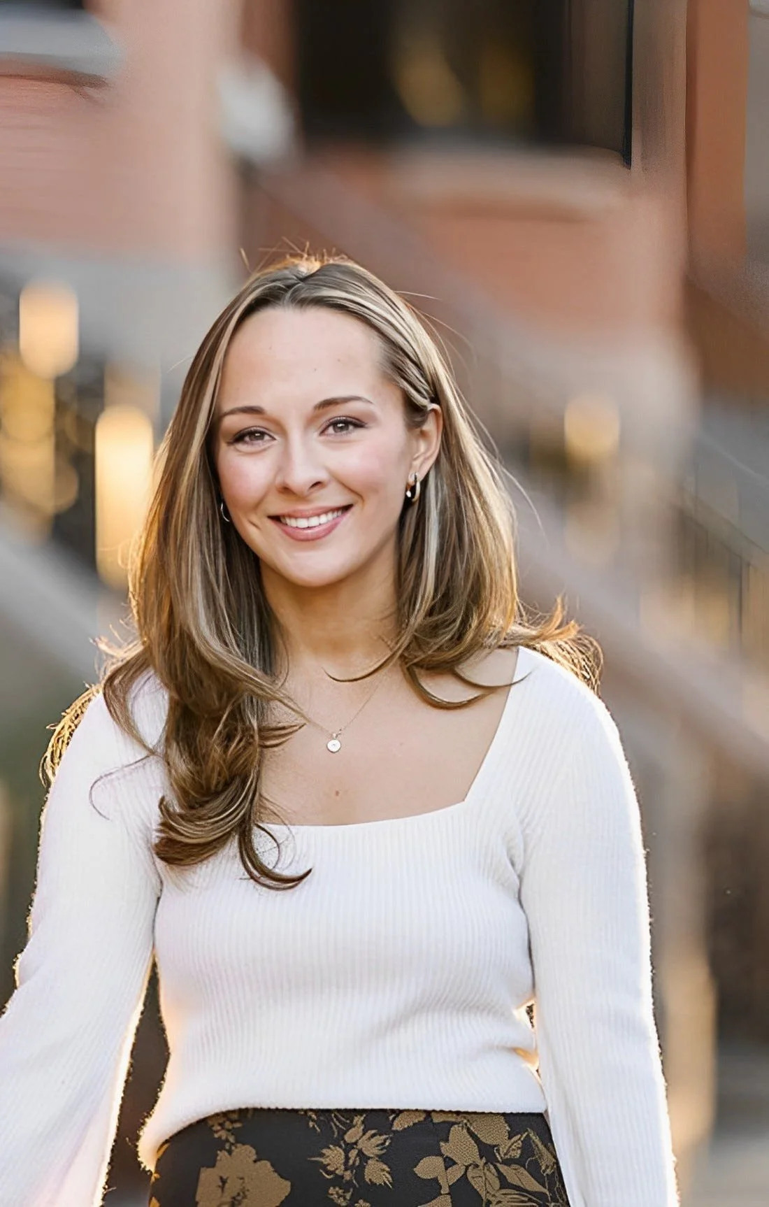 A young woman with long, wavy brown hair, smiling, wearing a white long-sleeve top and floral skirt, outdoors during sunset.