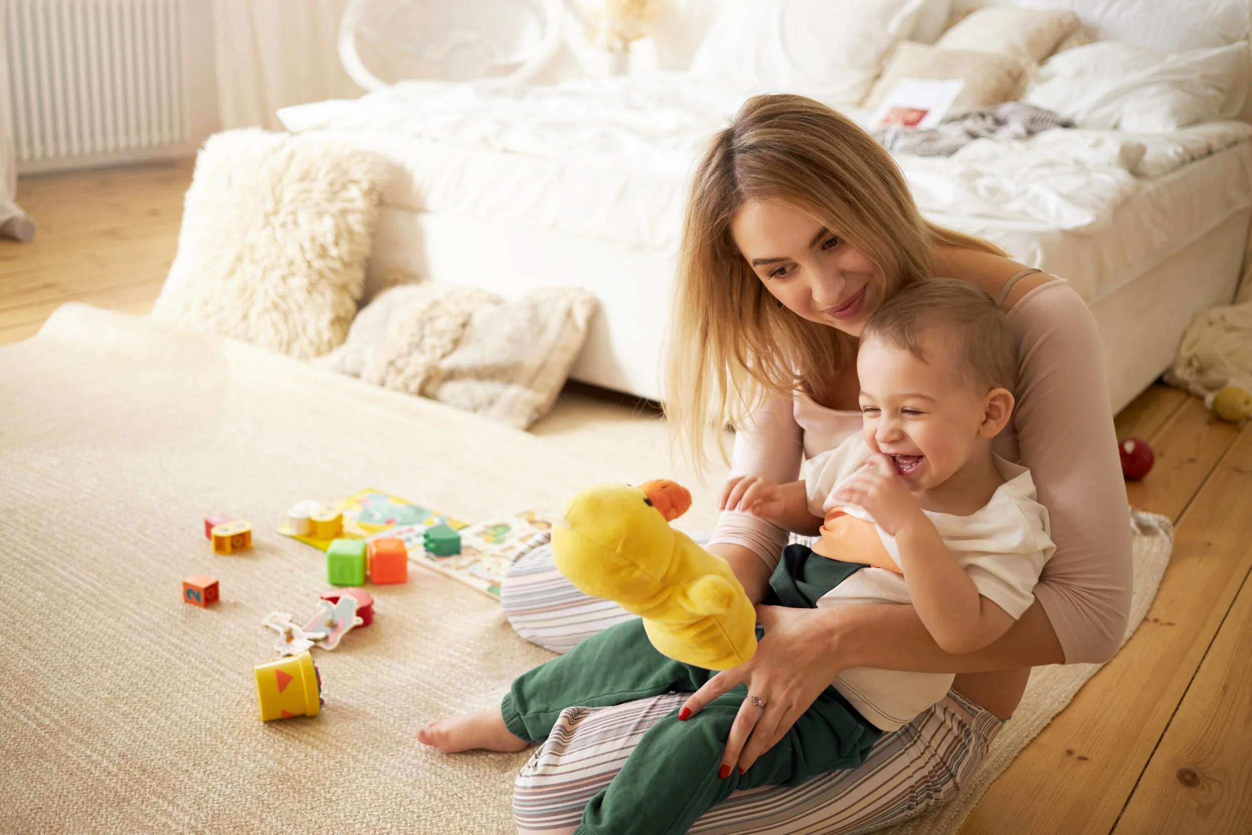 A woman and a young boy playing with a yellow plush duck toy on a beige carpet in a cozy bedroom. The bed in the background is unmade with pillows and blankets.