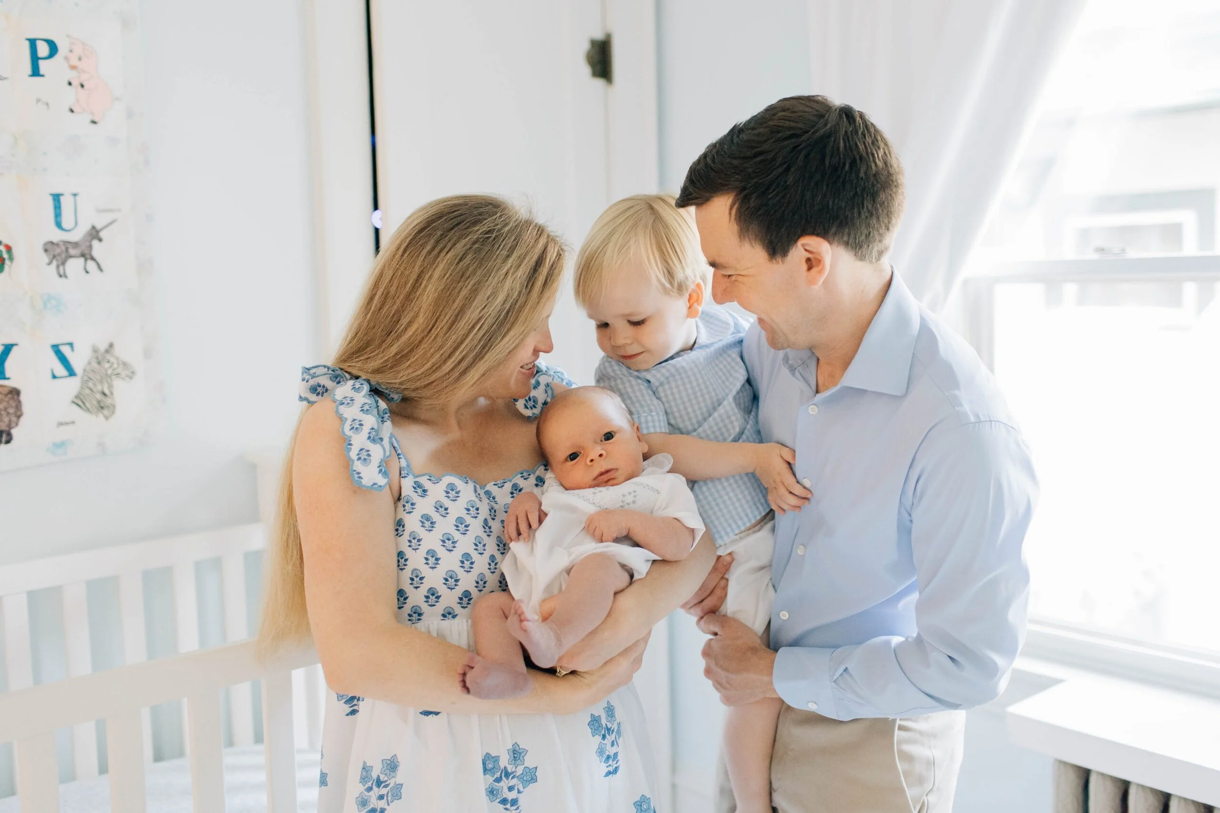 Family of four, including two young children and a baby, smiling and embracing indoors by a window.