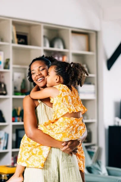 A woman holding a young girl in a yellow dress with white floral pattern, hugging and smiling in a bright room with a shelf in the background.