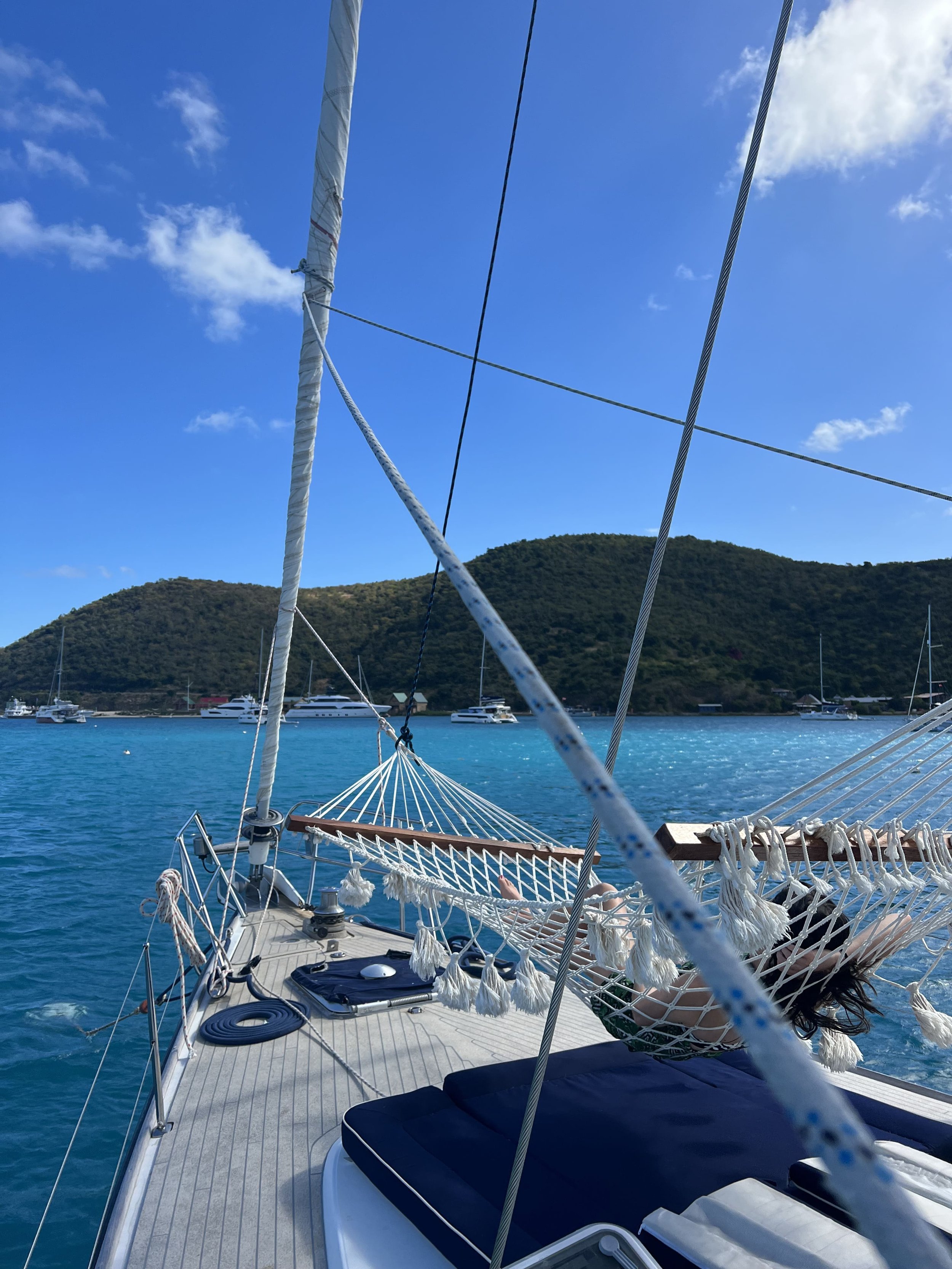 View of a boat's deck with a hammock, lounging area, and sailing rigging, overlooking a body of water with other boats and green, hilly islands in the background.