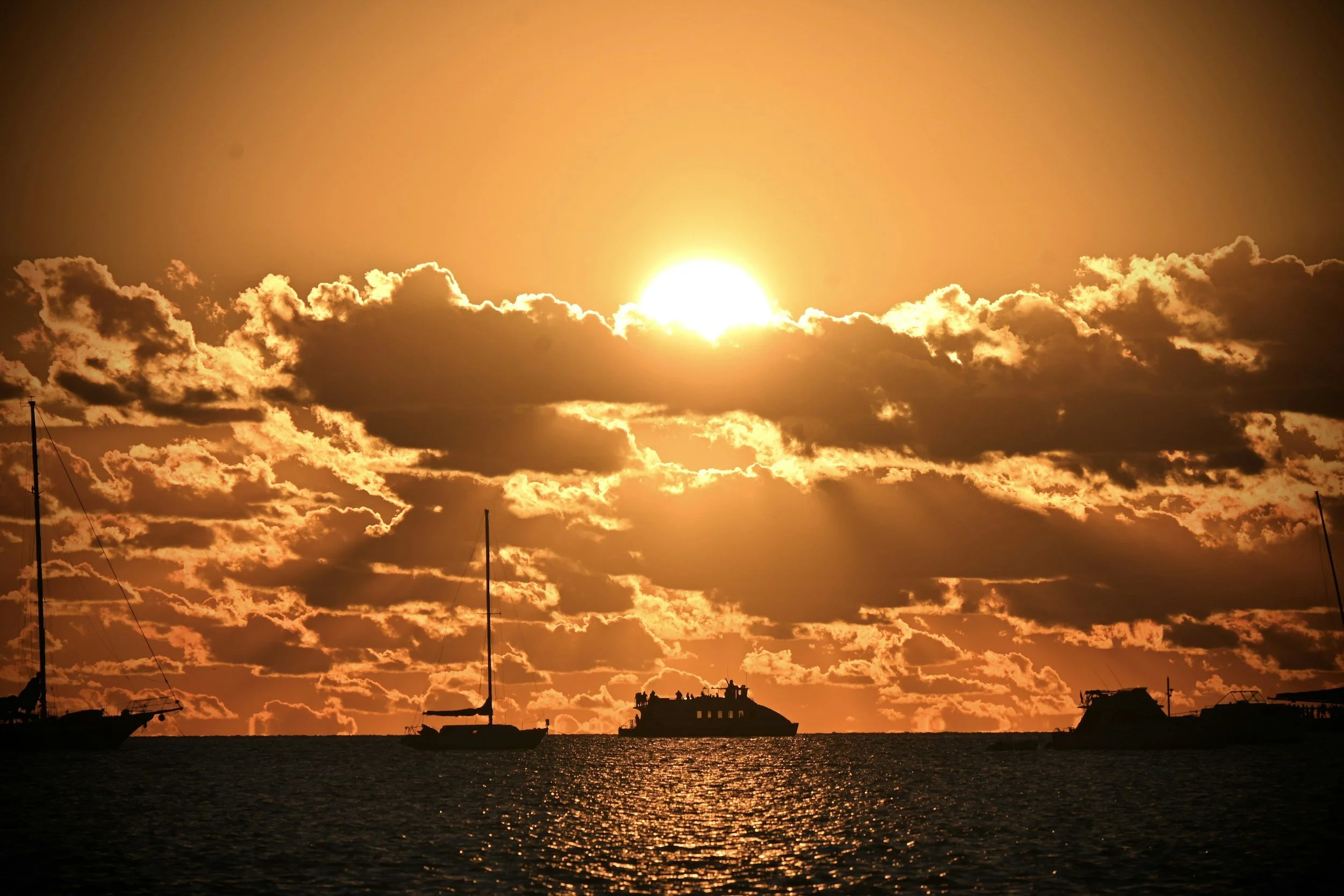 Sunset over the ocean with boats silhouetted against the sky, large clouds illuminated by the setting sun.