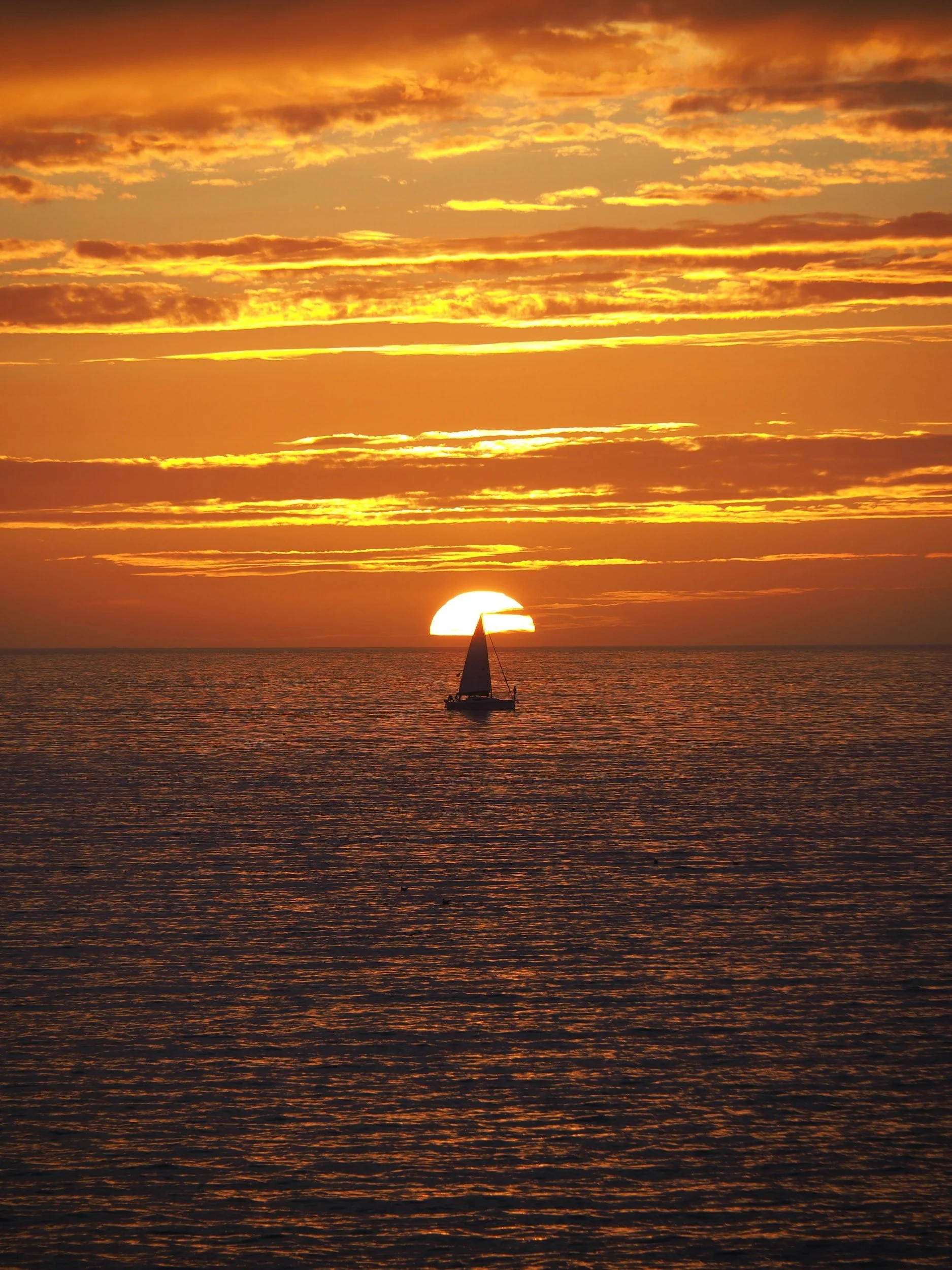 A sailboat on the ocean during sunset, with the sun partially below the horizon and a sky filled with orange and yellow clouds.
