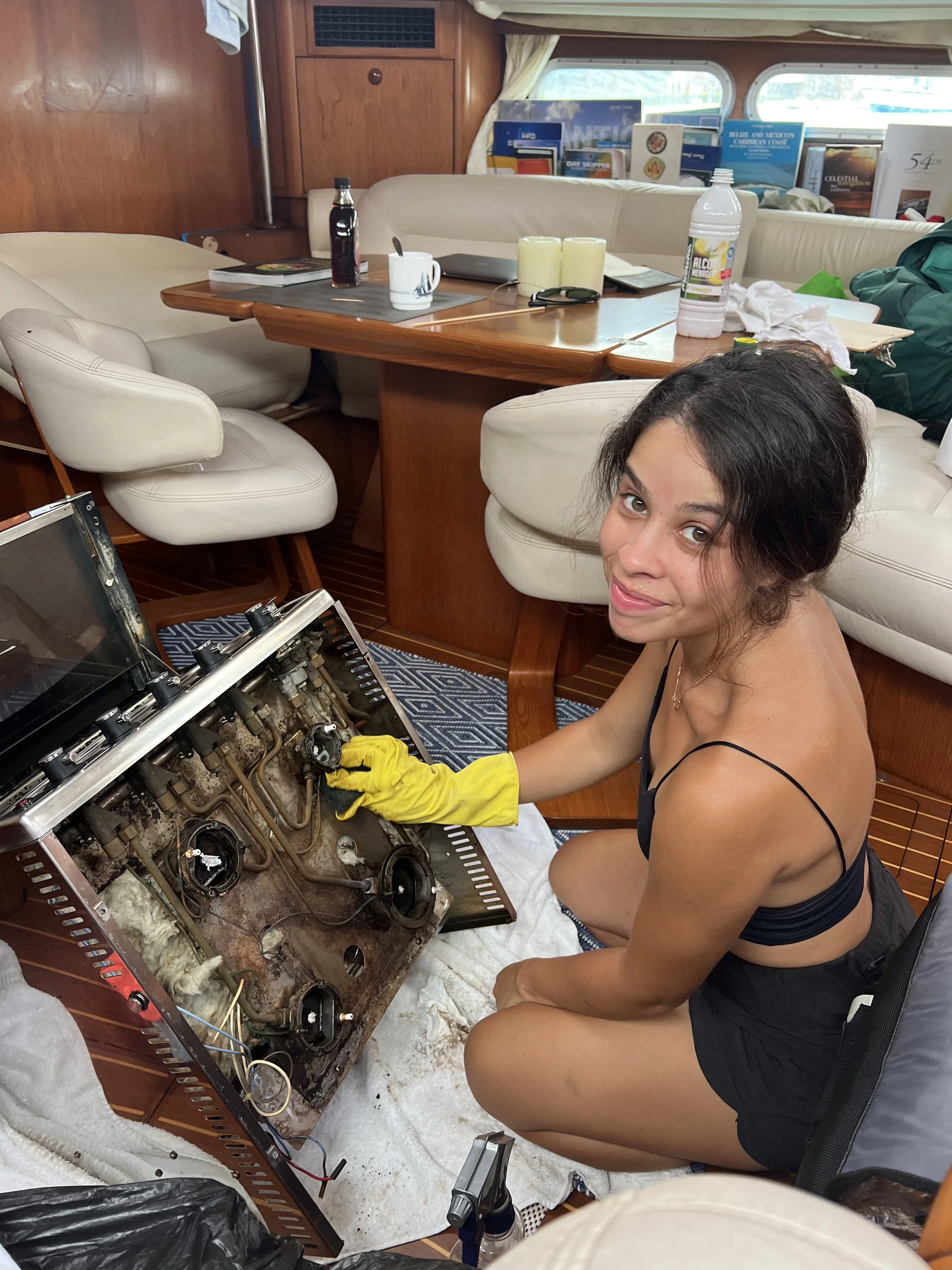 A woman cleaning or repairing a boat's engine inside a yacht. She is wearing yellow gloves, a black tank top, and shorts, and is crouching on a towel on the floor.