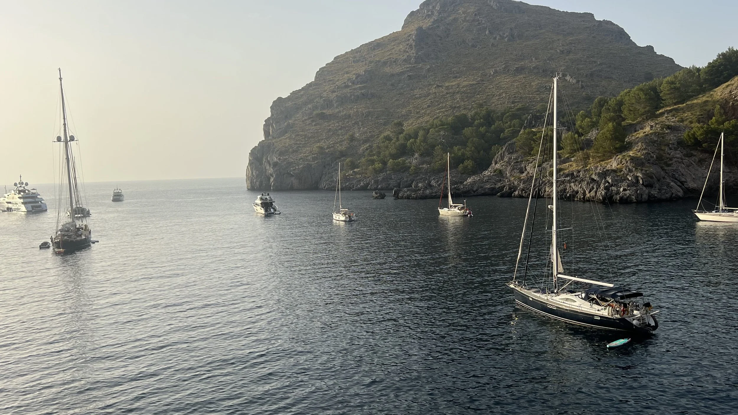 Multiple boats and yachts anchored near a rocky hillside in calm water, with a hazy sky.