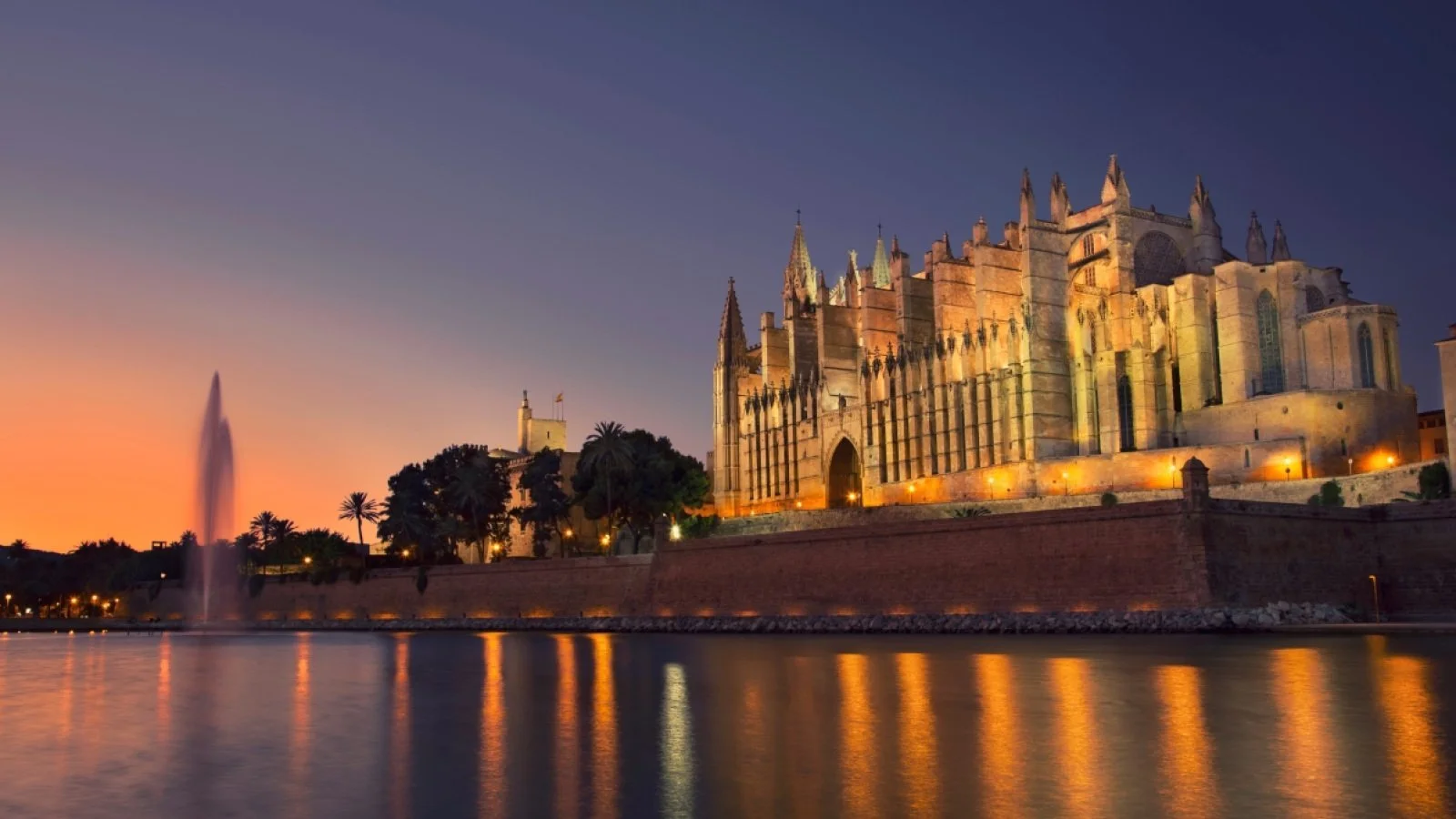 Historic cathedral by the water at sunset with a fountain in the foreground and palm trees. The sky is a gradient of orange and deep blue.