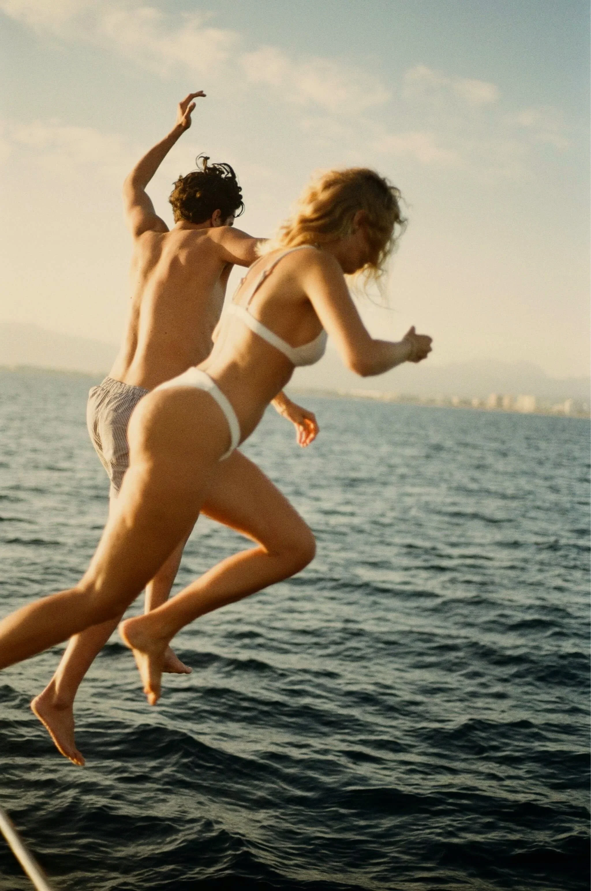 Two people in swimsuits jumping into the water off a boat on a sunny day, with sea and distant skyline in the background.
