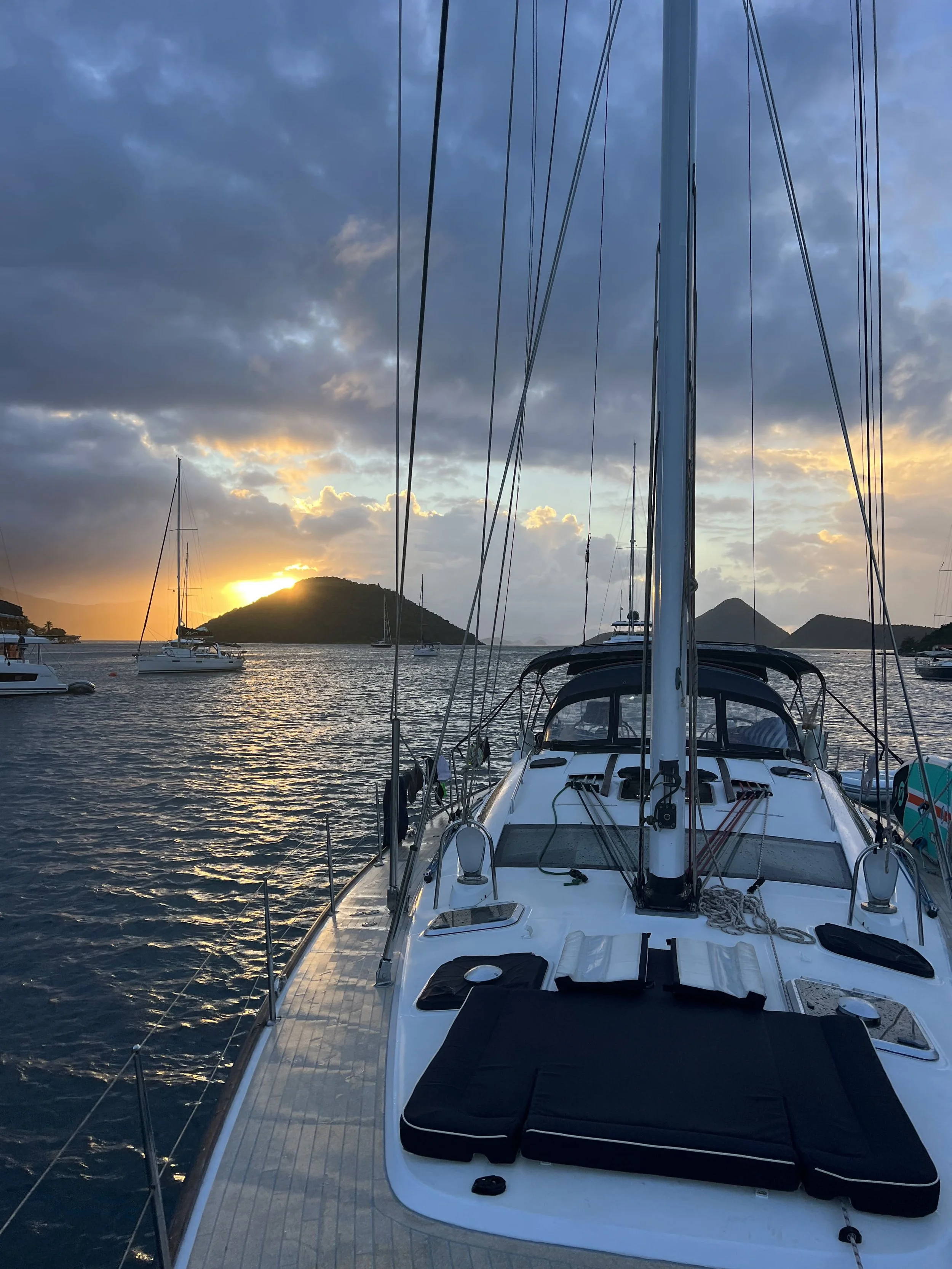 View from a yacht at sunset over a calm bay with other sailboats anchored nearby and a hill in the background.