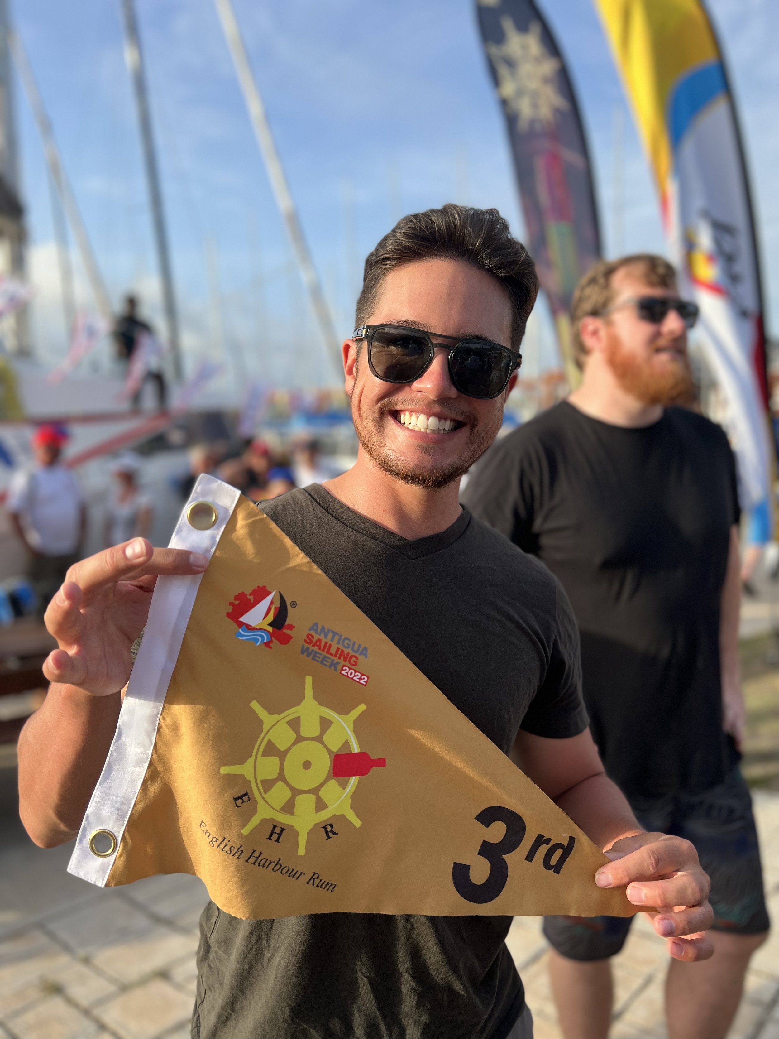 A smiling man wearing sunglasses holding a yellow racing flag with '3rd' written on it, at an outdoor event with boats and flags in the background.