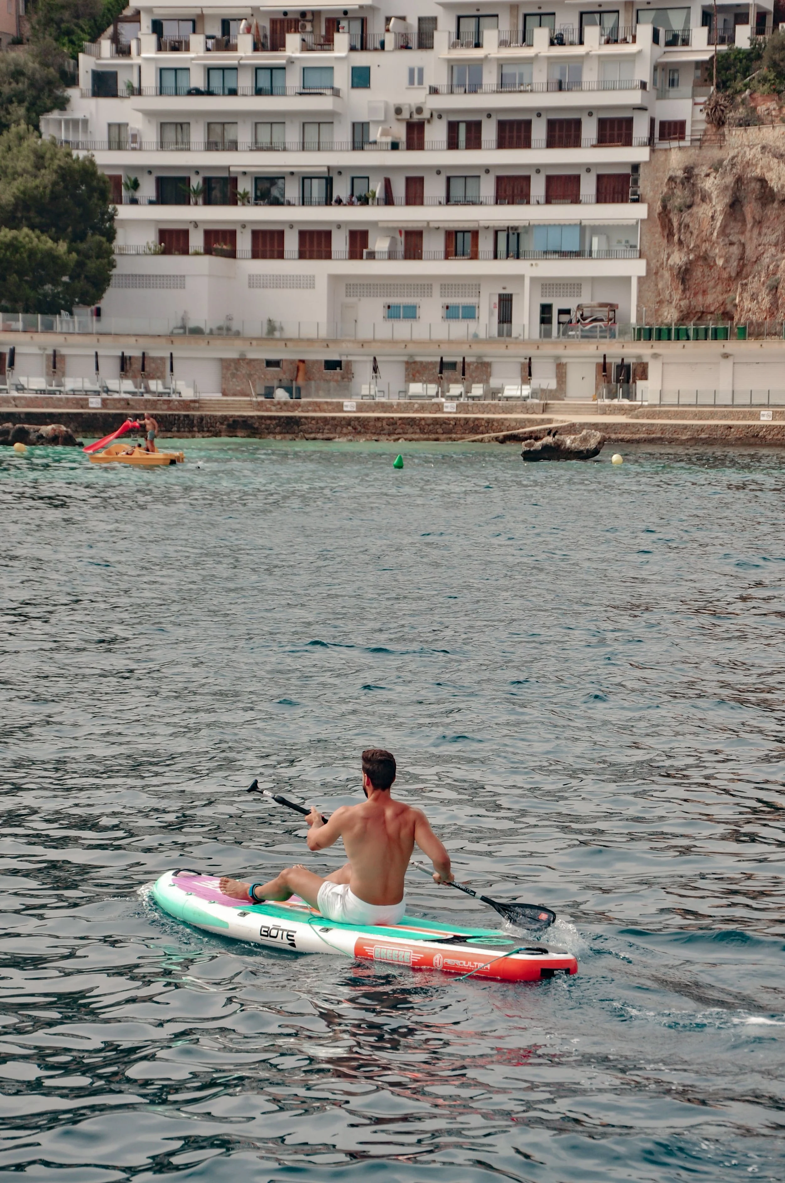 A man in swim shorts paddleboarding on a calm body of water with a white and green stand-up paddleboard, near a shoreline with a multi-story hotel or apartment building in the background.
