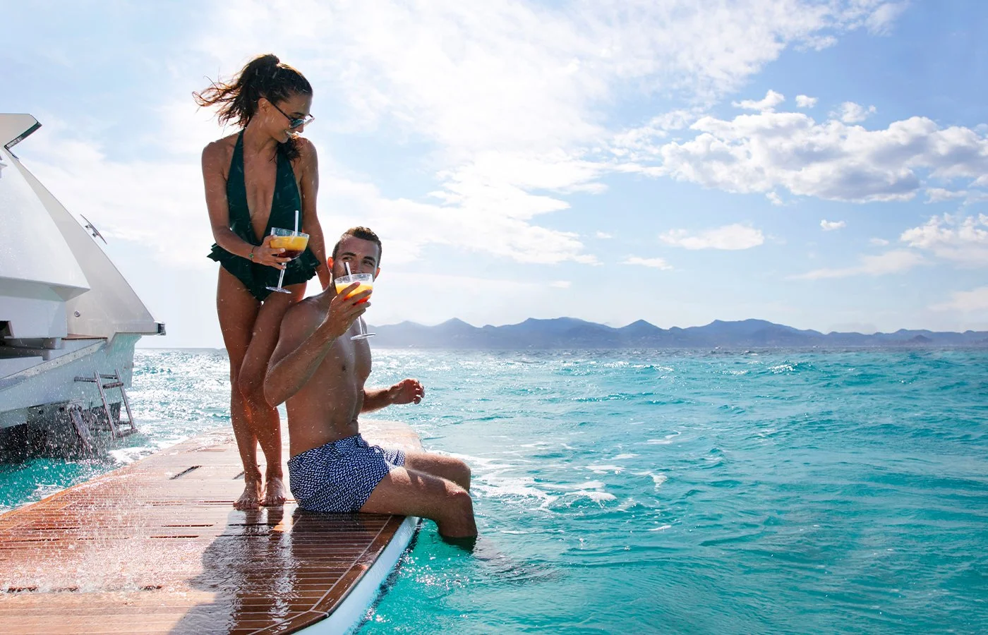 couple having drinks on a boat swimming platform on crystal clear waters and mountains in the brackground on a sunny day