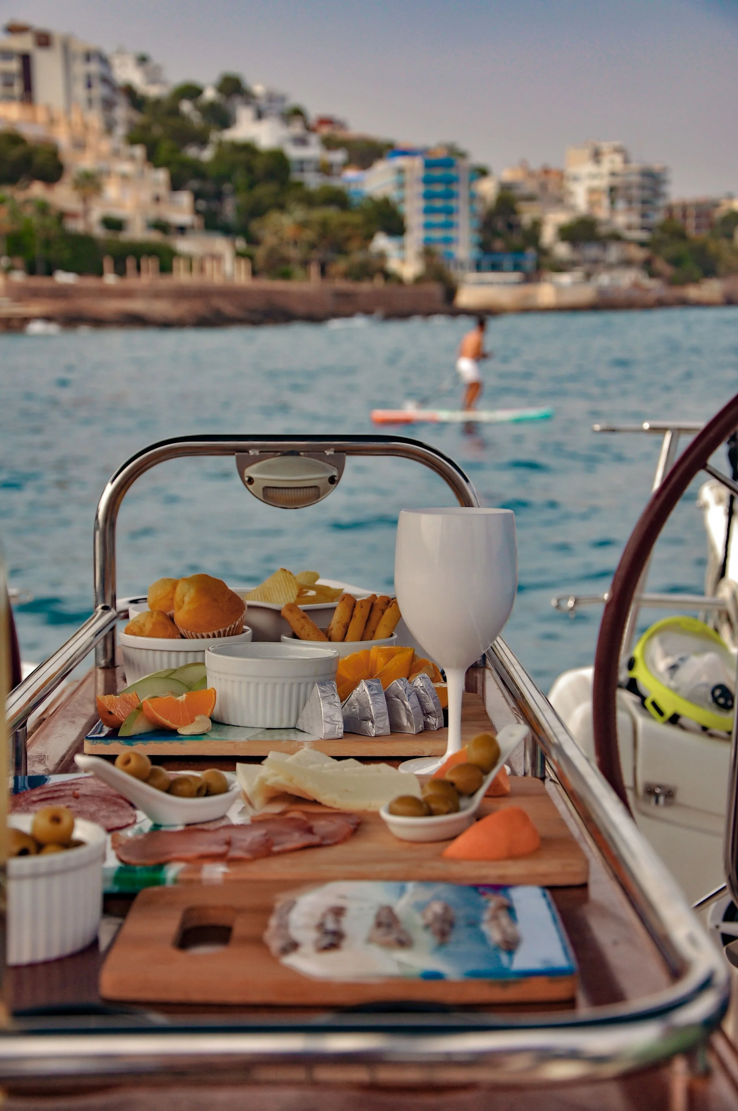 A boat's table with assorted food and drinks on a deck, overlooking water with a paddleboarder in the background and a cityscape on the shoreline.