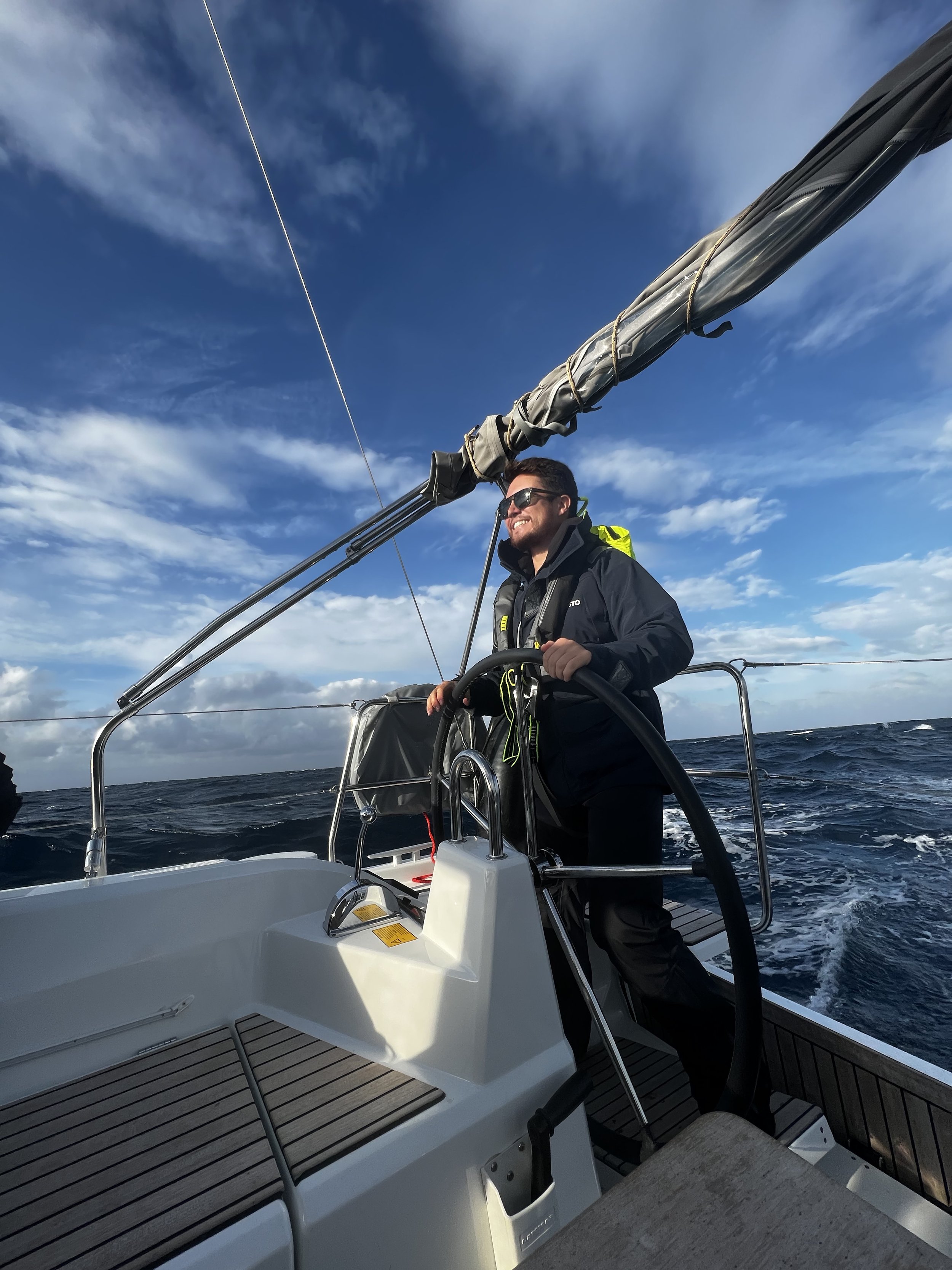 Man with sunglasses sailing a boat on the ocean during daytime. He is standing at the helm, holding the steering wheel, with a bright blue sky and some clouds overhead.