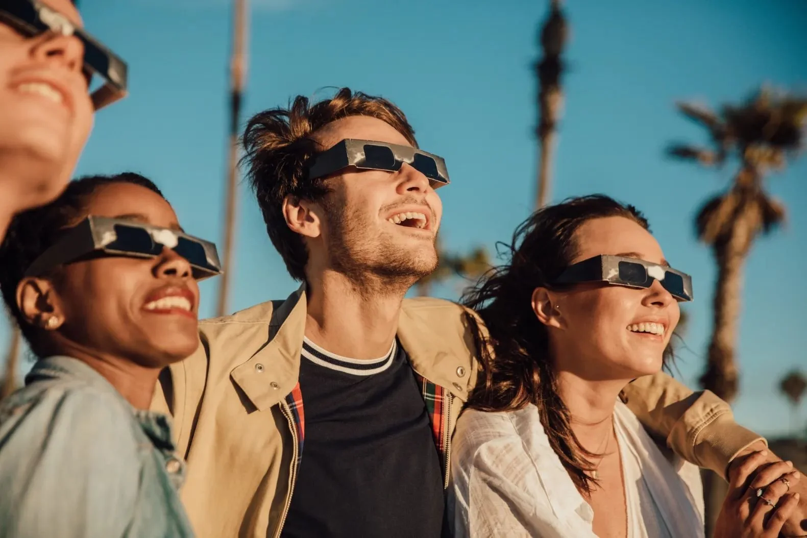 Group of four diverse friends wearing solar eclipse glasses, smiling and enjoying together outdoors on a sunny day with palm trees in the background.