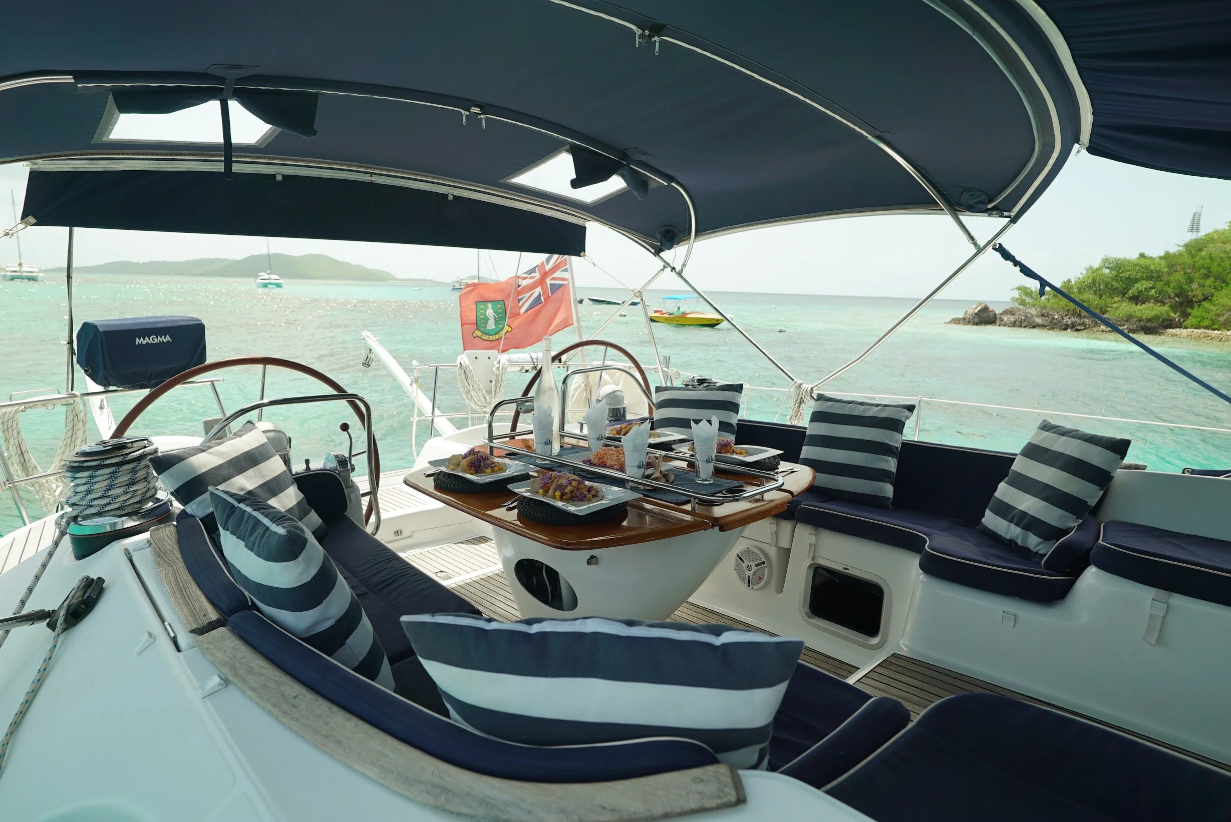 A yacht with a shaded deck, striped cushions, and a table set with food and drinks, anchored near a green island with a calm, turquoise sea.