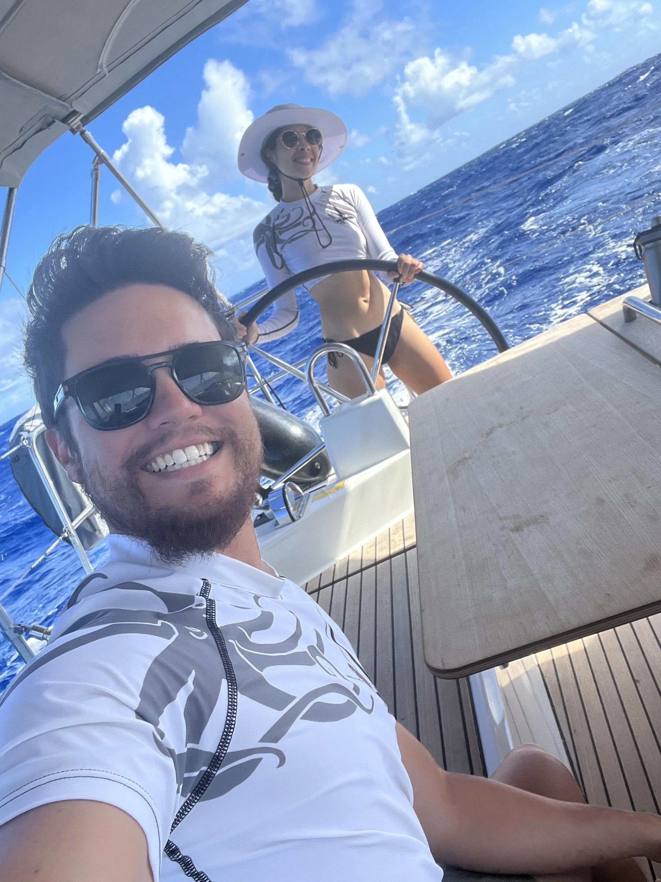 A smiling man with sunglasses taking a selfie on a boat with a woman steering in the background, both enjoying a sunny day on the ocean.